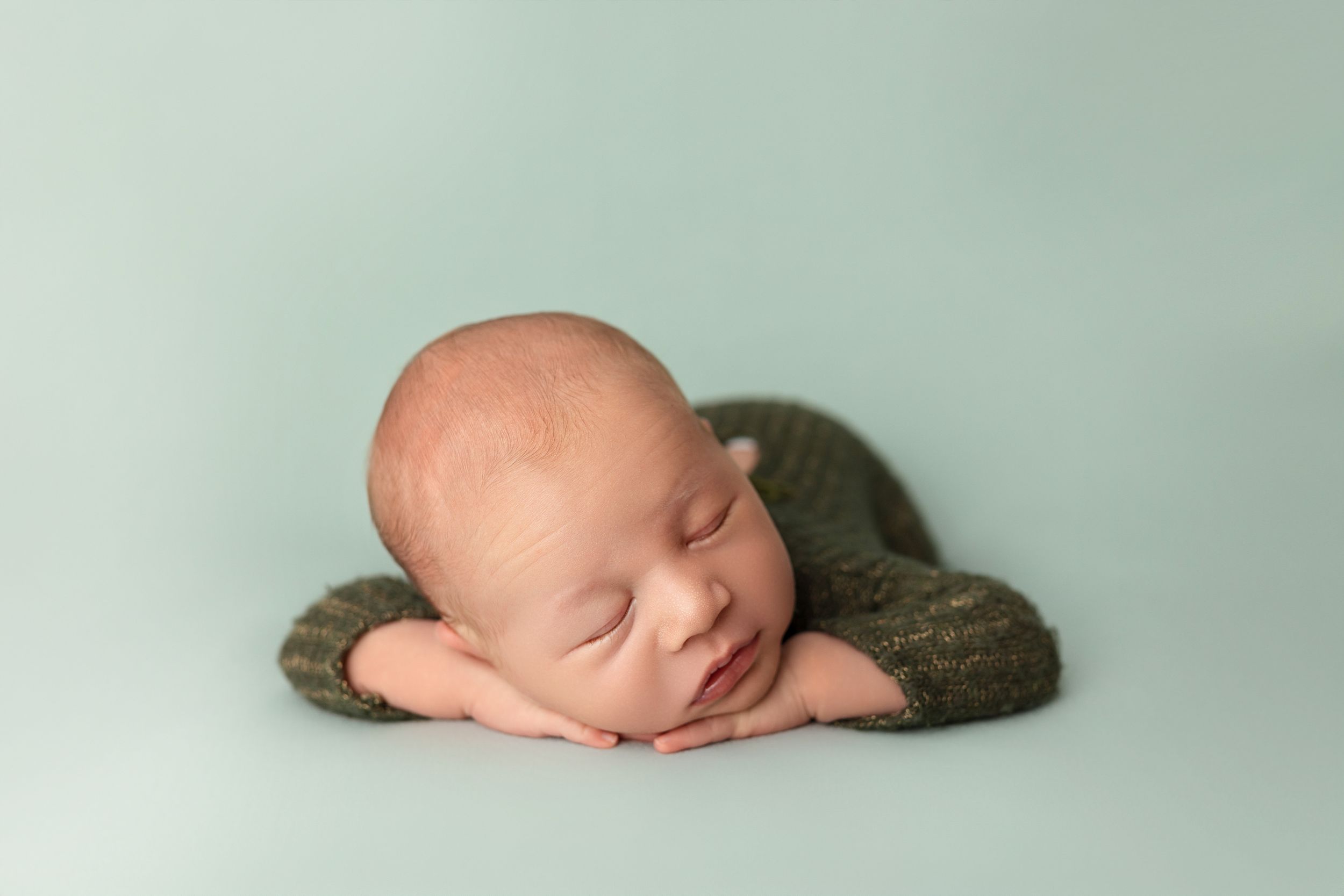 A sleeping newborn rests peacefully on a mint green backdrop with arms folded under their head.