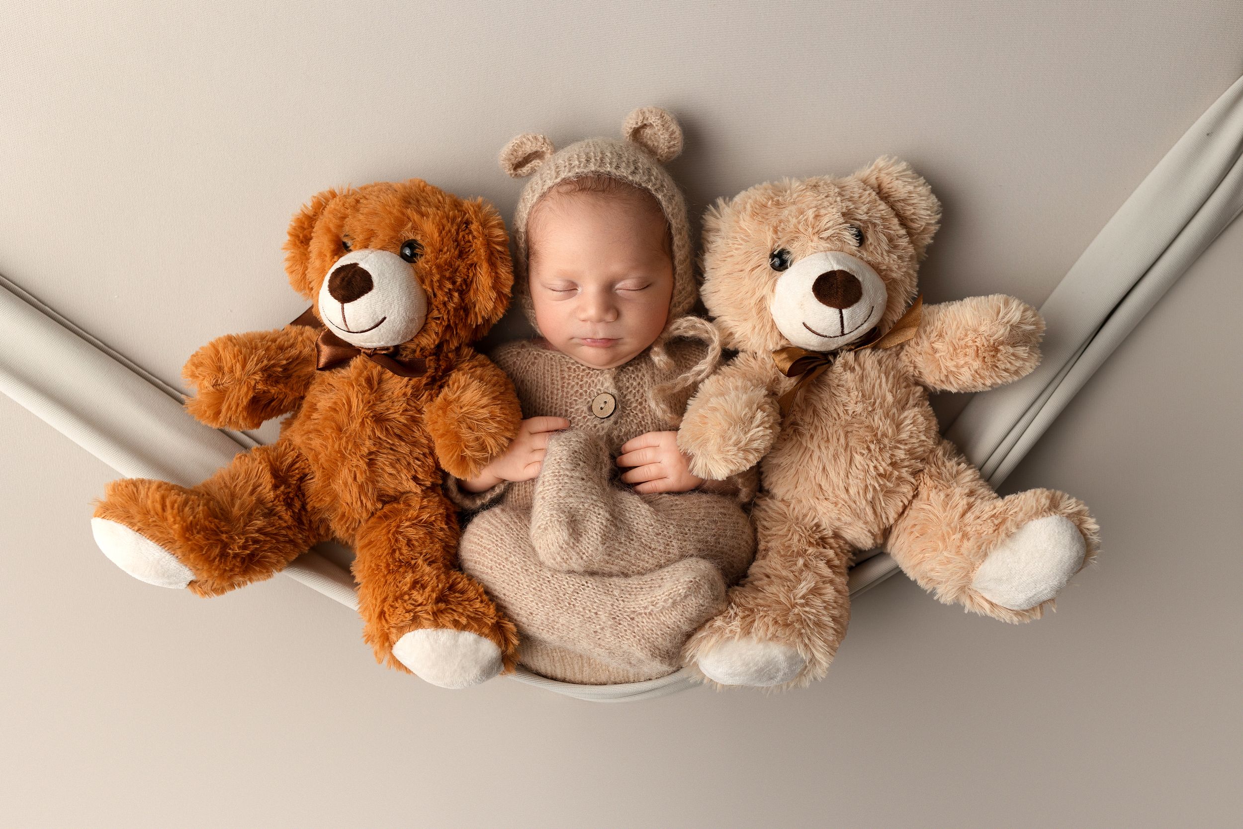 Sleeping baby nestled between two plush teddy bears, a brown and beige one, on a neutral backdrop.