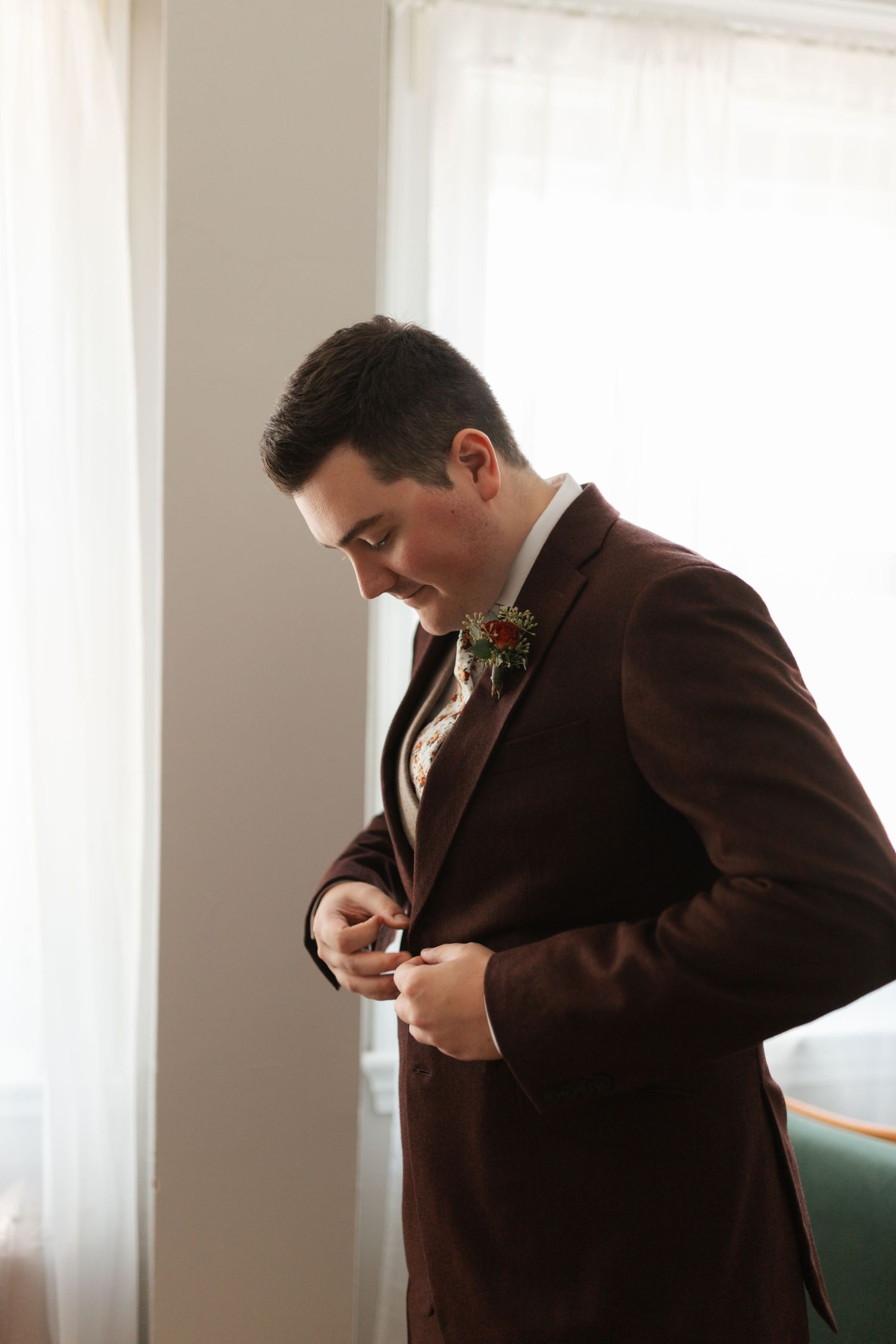 Groom in burgundy suit adjusting cufflinks before wedding ceremony near bright window.