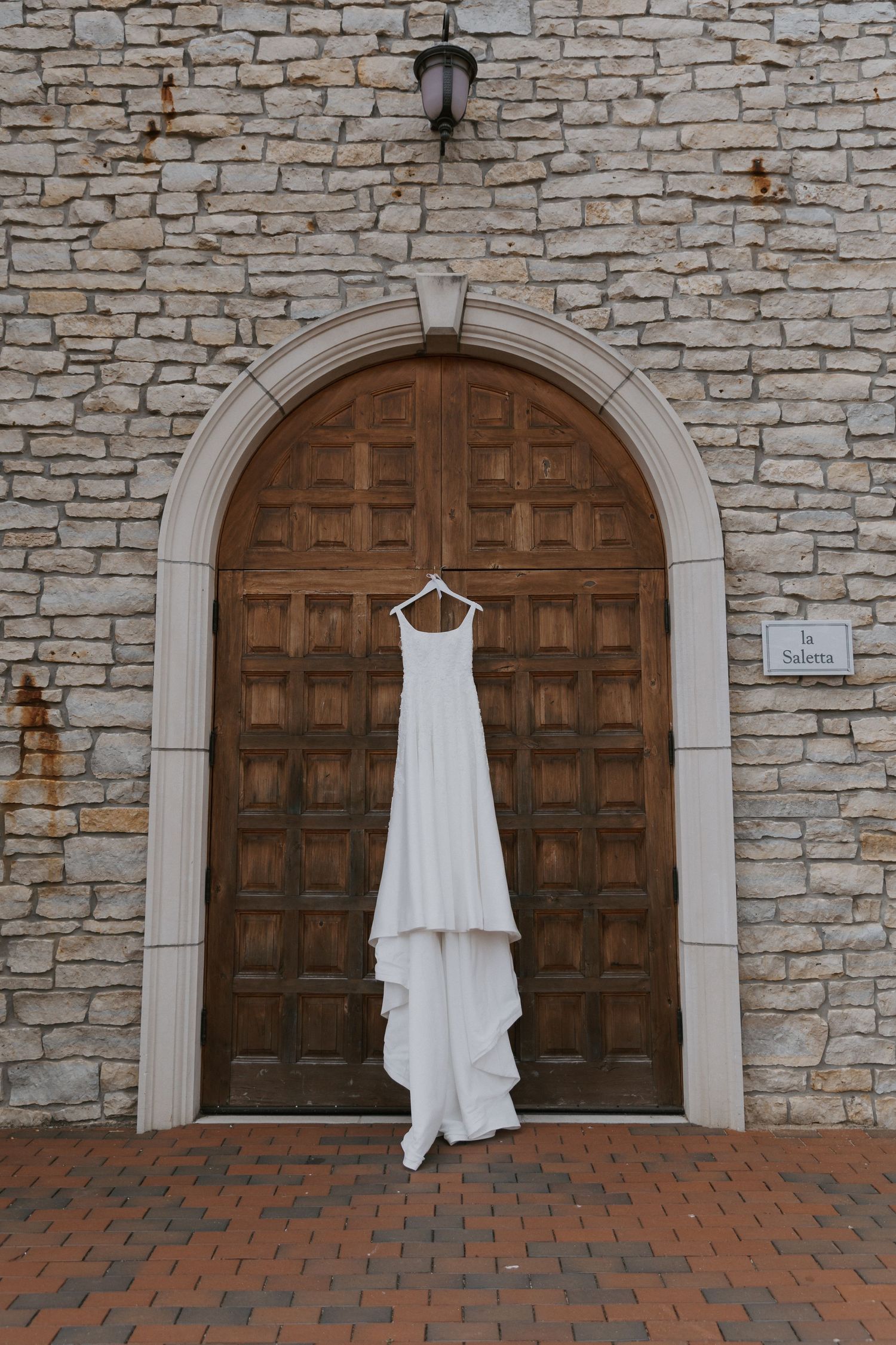 A white wedding dress hangs centered on large wooden arched door against stone wall in the piazza cocktail hour area.