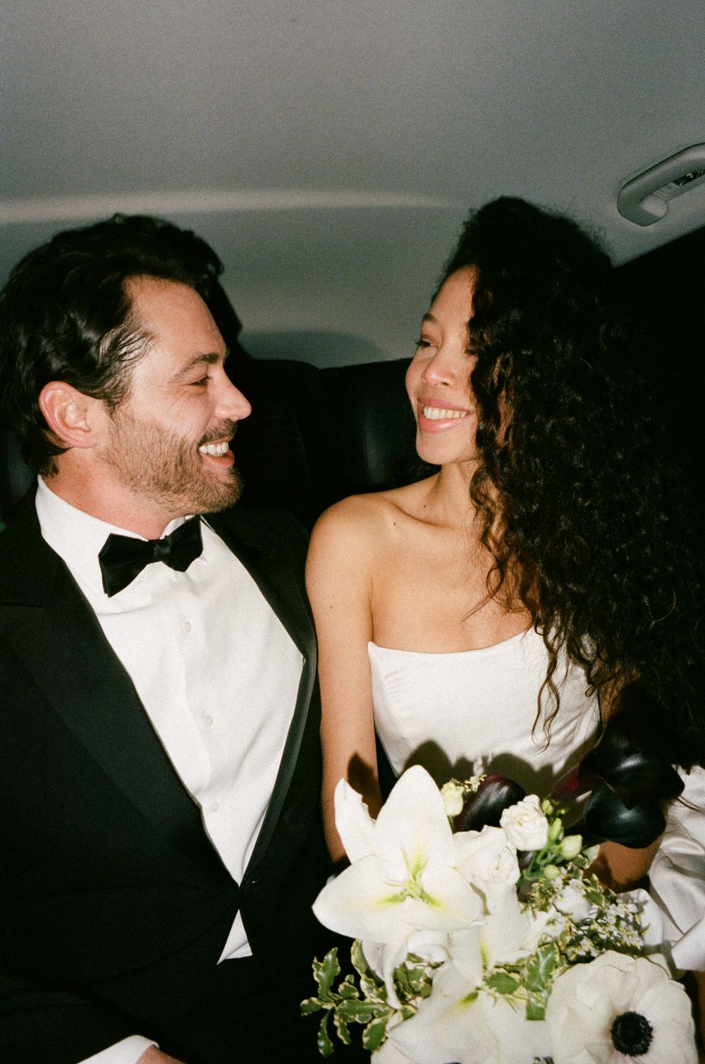 A happy couple smiling at each other in formalwear while holding a bouquet of white flowers in what appears to be a car.