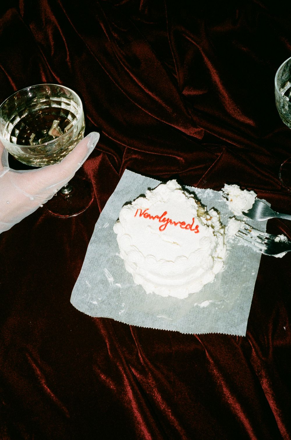 A small white frosted cake with red writing sits beside a glass of wine on a dark velvet surface.