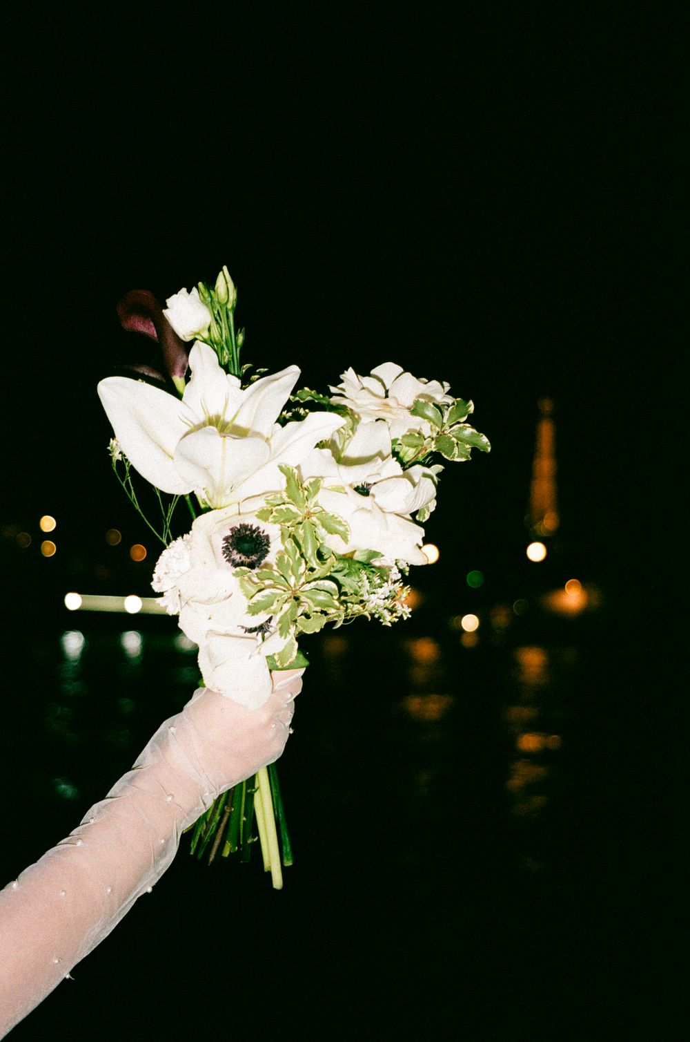 A hand holds a delicate white floral bouquet with lilies at night against a dark background with bokeh lights.