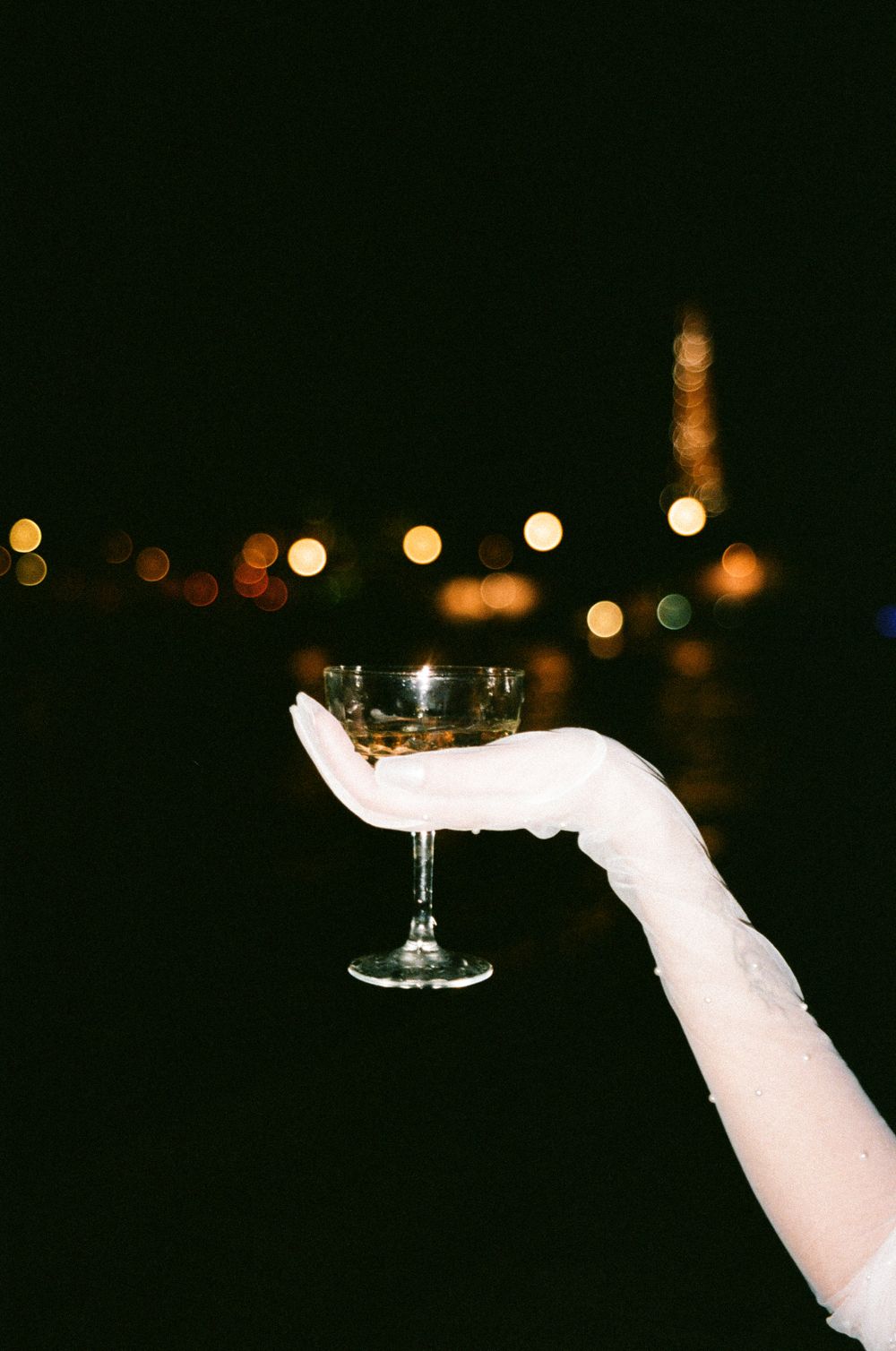 A hand in white glove holds a cocktail glass against a dark background with bokeh lights.