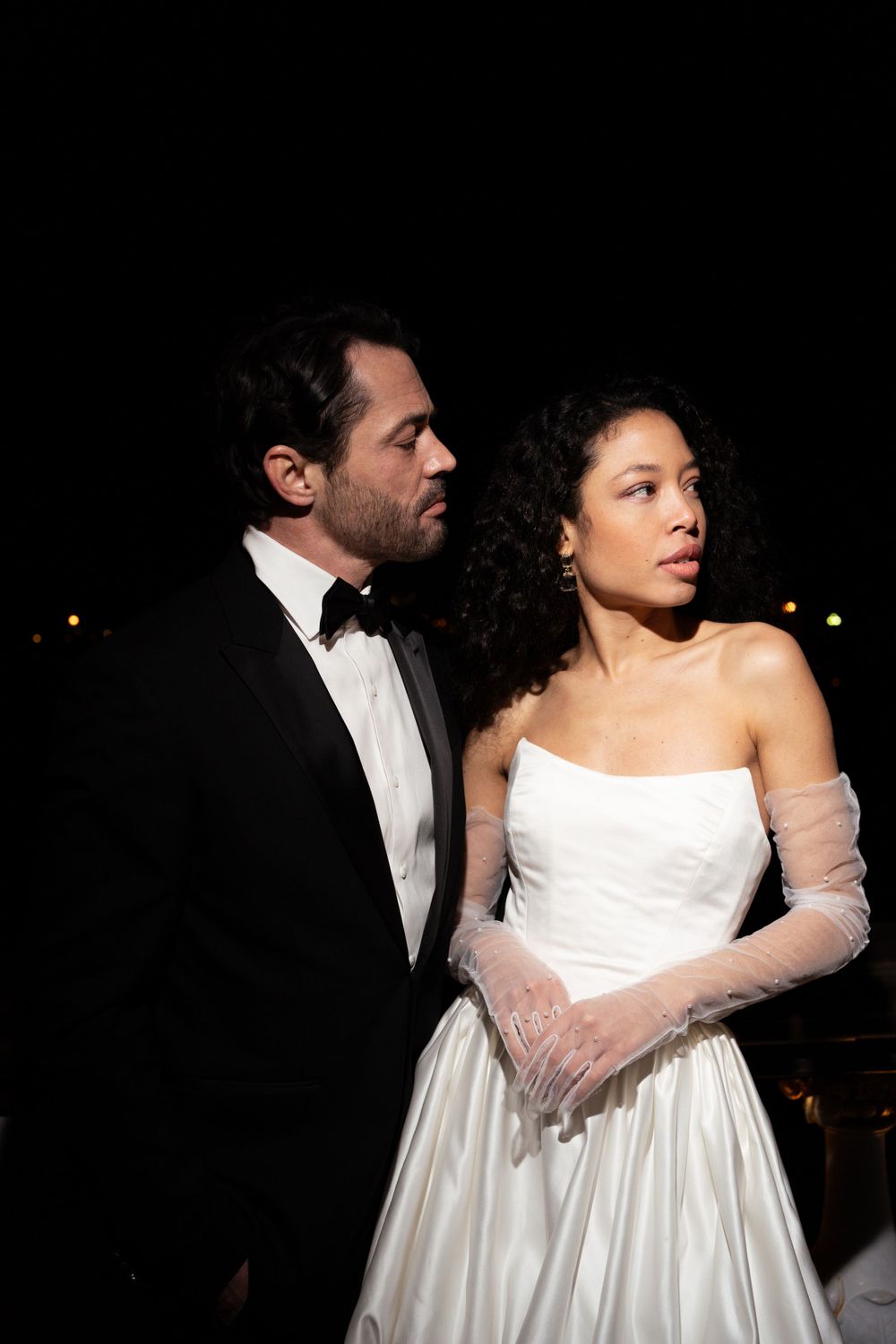A couple in formal wedding attire stands together at night, with the bride in a white gown and long gloves.