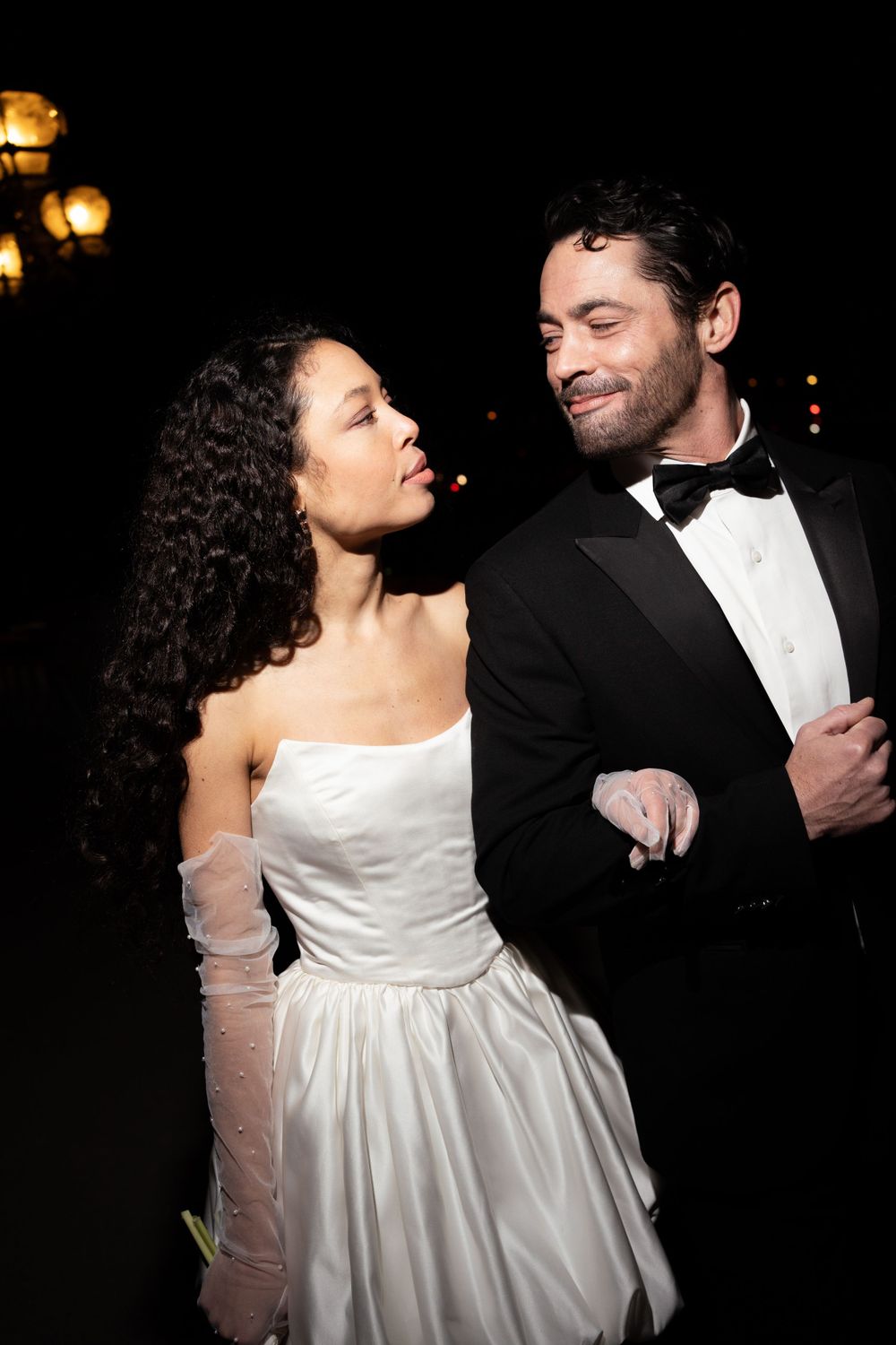 A couple in formal wedding attire share a romantic moment at night, with the bride in white and groom in a tuxedo.