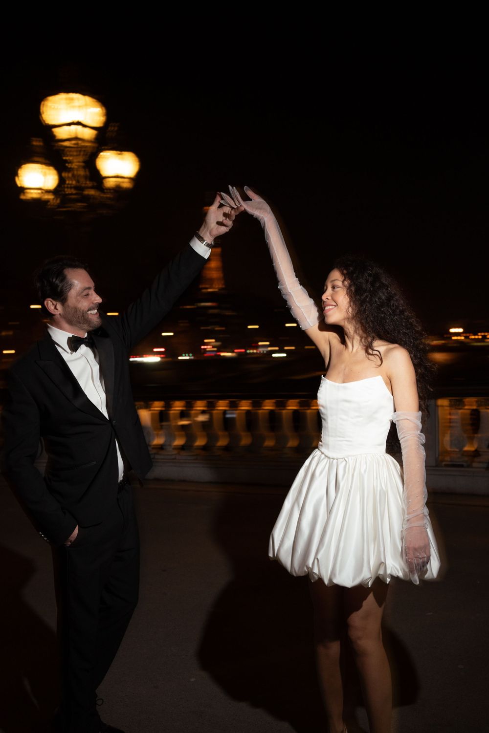A couple in formal attire dancing together at night under illuminated lamps on a bridge.
