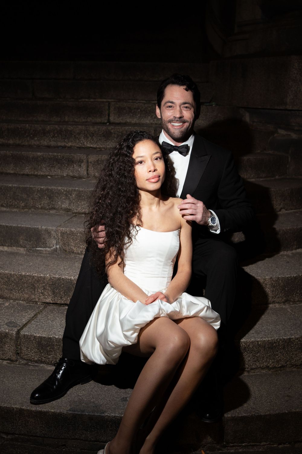 A couple in formal attire sit on stone steps, with the bride wearing a white dress and groom in classic black tuxedo.