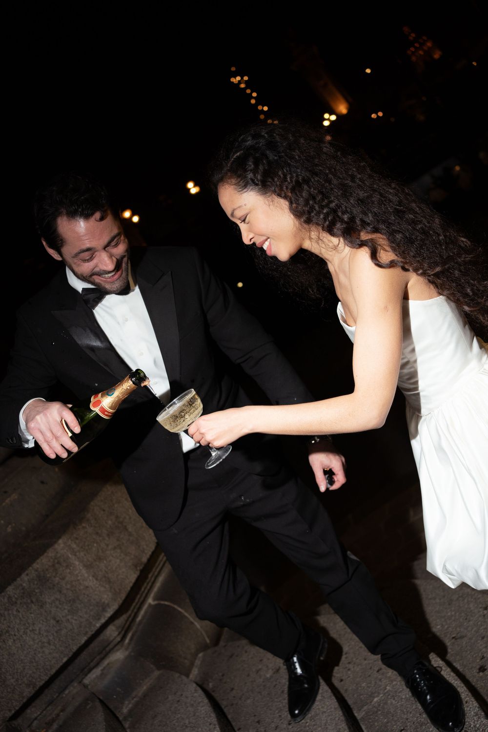 Two elegantly dressed wedding guests laugh and dance together while holding champagne glasses at a nighttime celebration.
