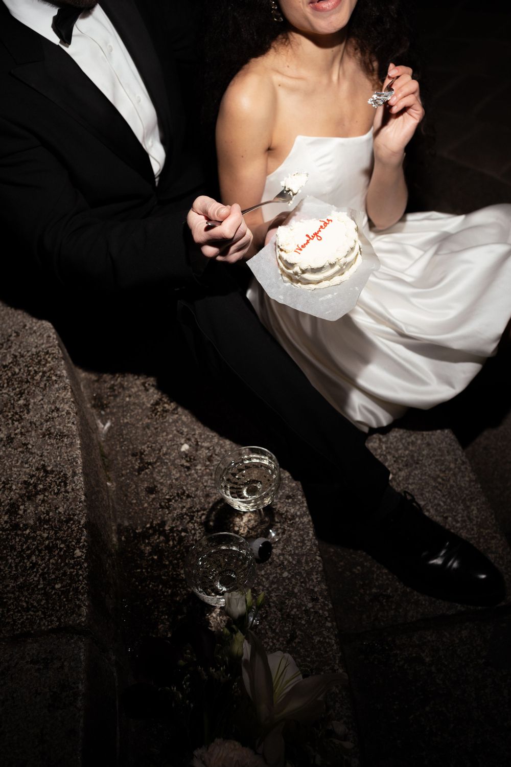 A couple in formal attire sits together while enjoying wedding cake, with champagne glasses reflective on dark ground.