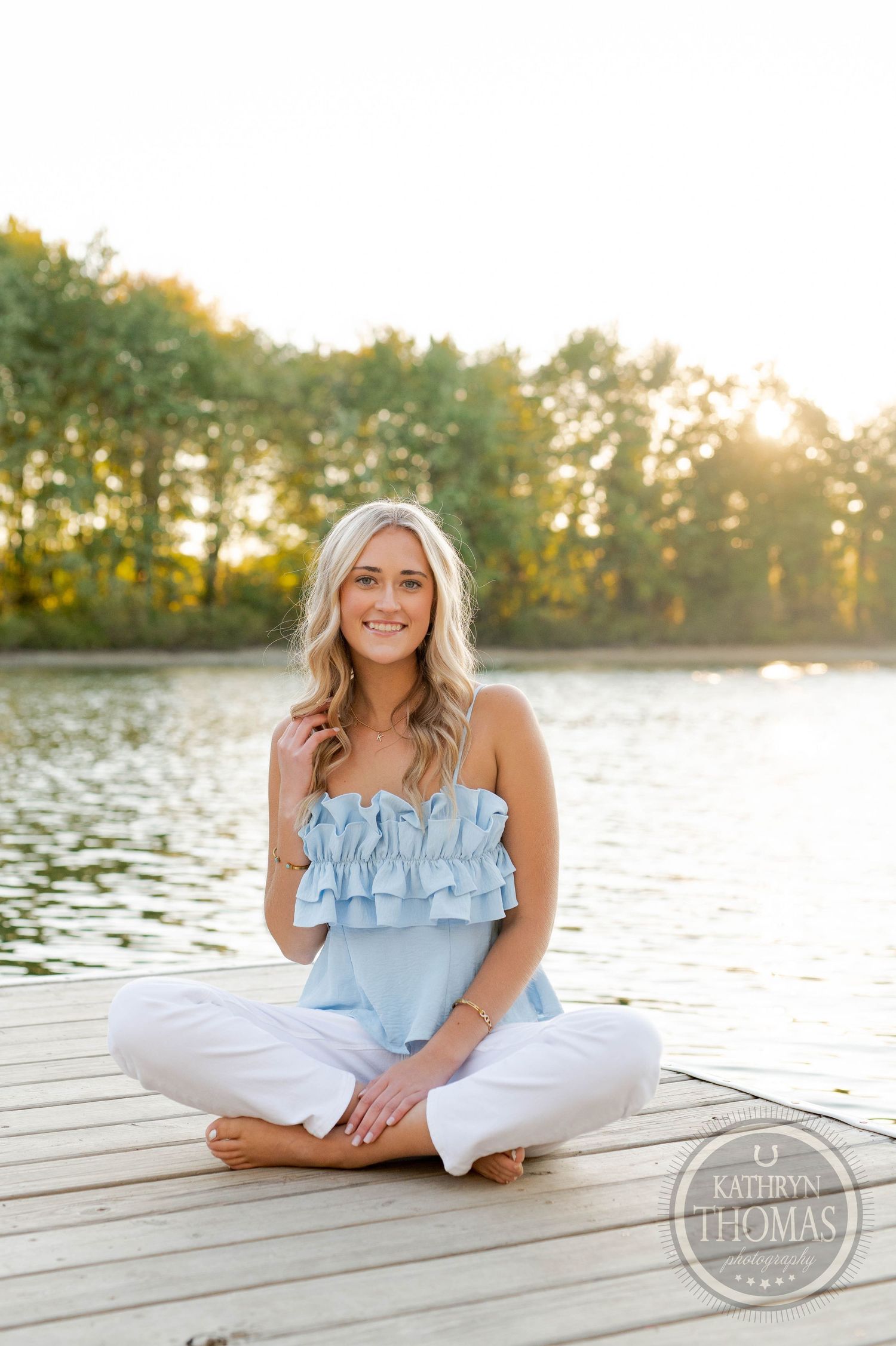 Person in white pants and blue ruffled top sitting peacefully on wooden dock during sunset.