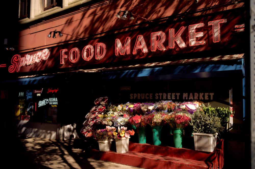 Vintage food market storefront with flower displays under red awning.