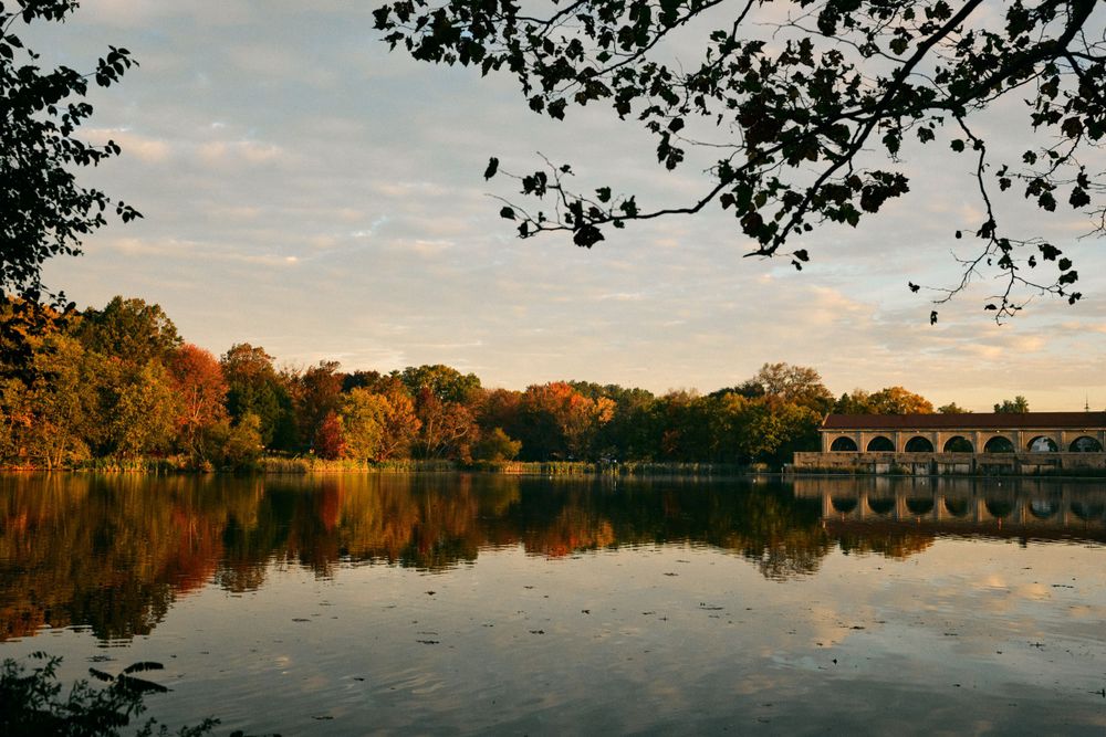 Stone bridge reflects in calm lake water during golden sunset with autumn trees.
