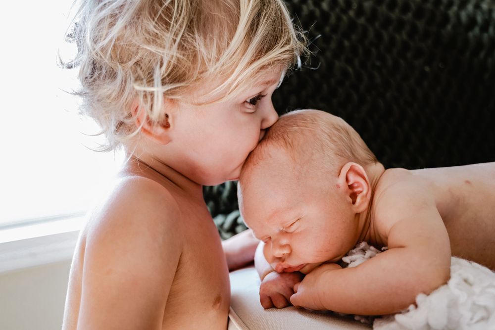 Young child giving sleeping newborn sibling gentle kiss on head.