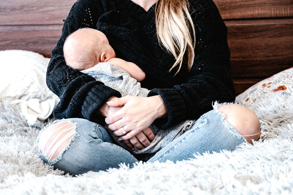 Someone in a black sweater and ripped jeans sitting on white carpet holding a small infant.