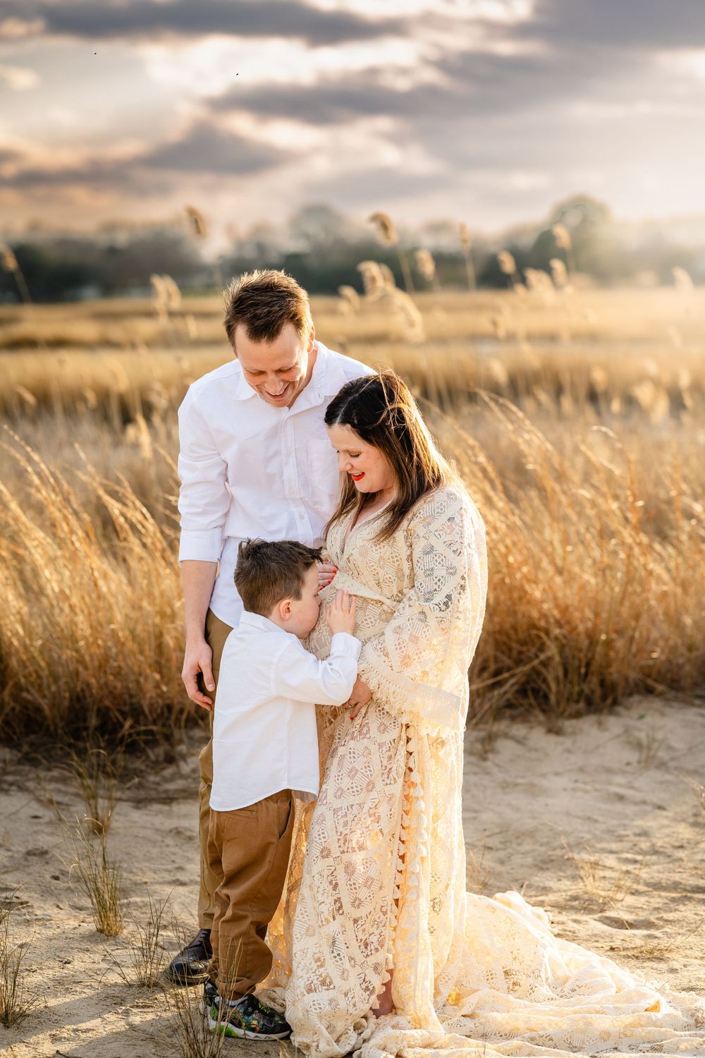 A family in white and cream colored outfits sharing a tender moment in a golden field at sunset.