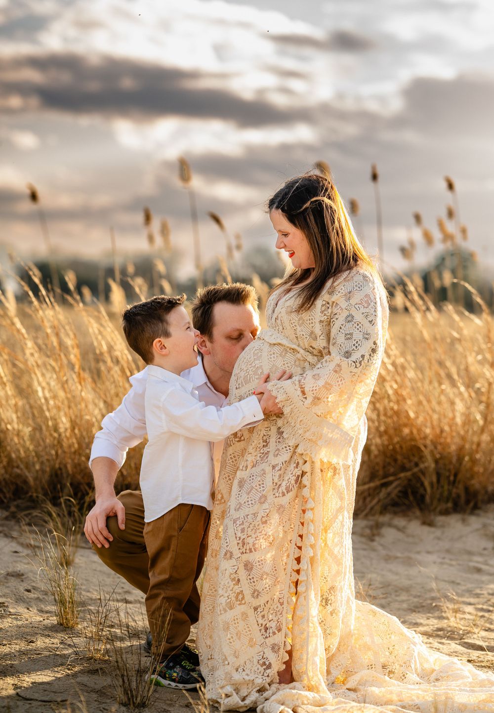 A family shares an intimate moment in a golden wheat field at sunset, wearing white and cream-colored outfits.