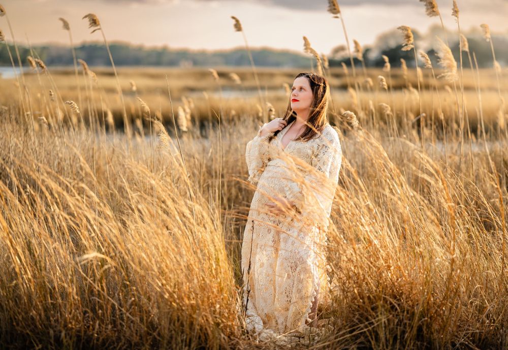 A woman in a white lace dress stands among golden reeds in a marsh at sunset.