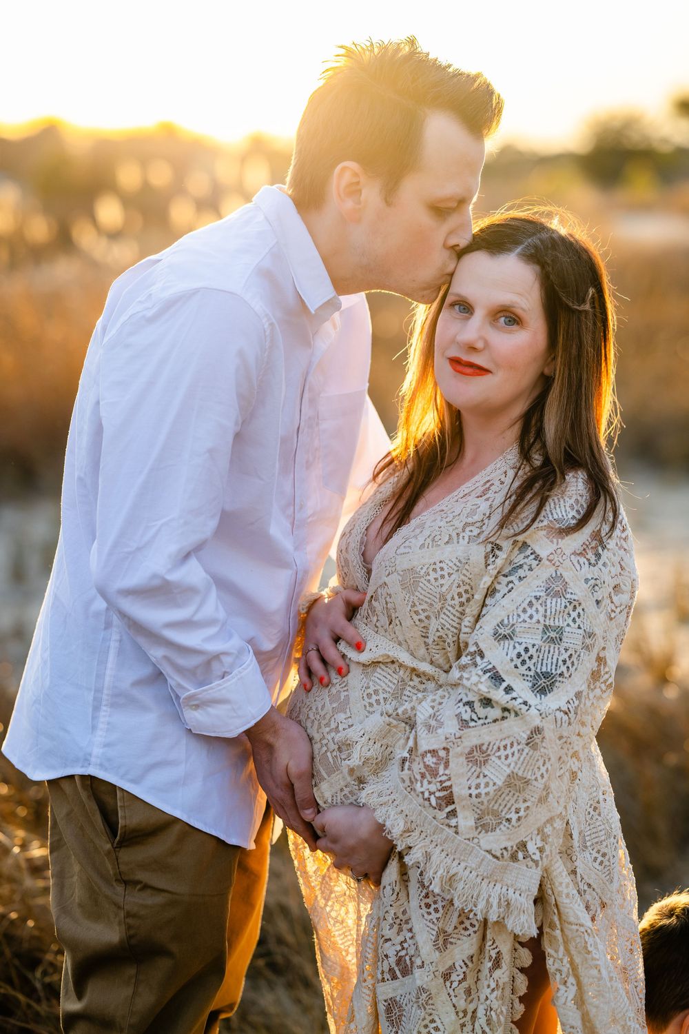 A couple poses in a sunlit field with glowing backlight during a maternity photo session at golden hour.