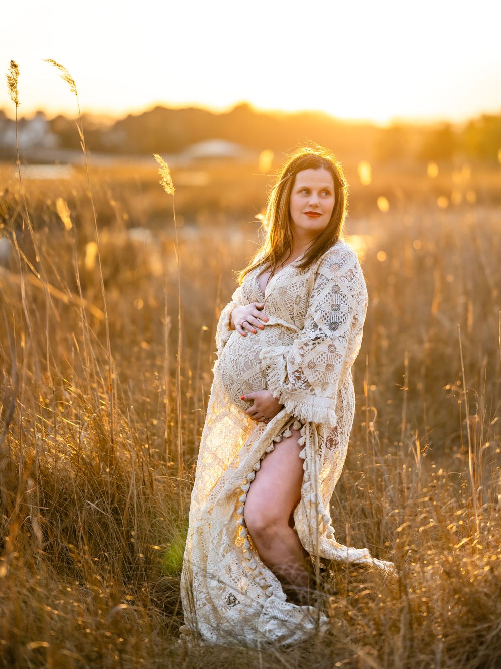 A pregnant figure in a white lace dress poses in a golden field at sunset creating a dreamy maternity portrait.