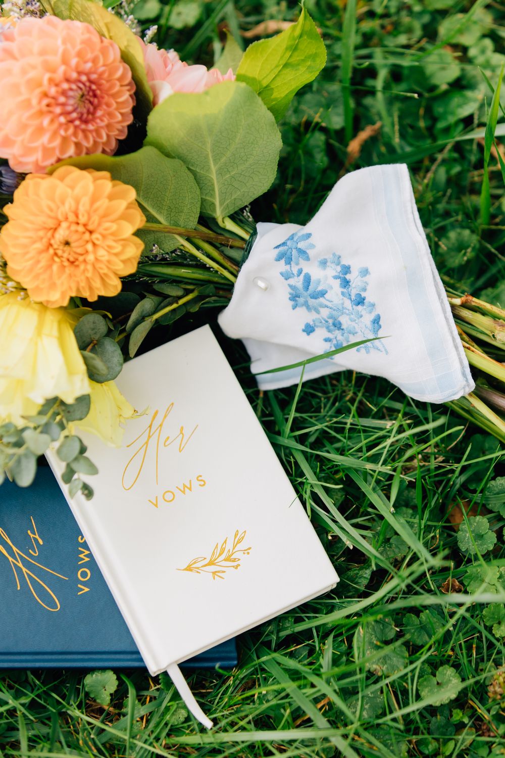 Wedding vow books with gold lettering lay among peach dahlias on grass.