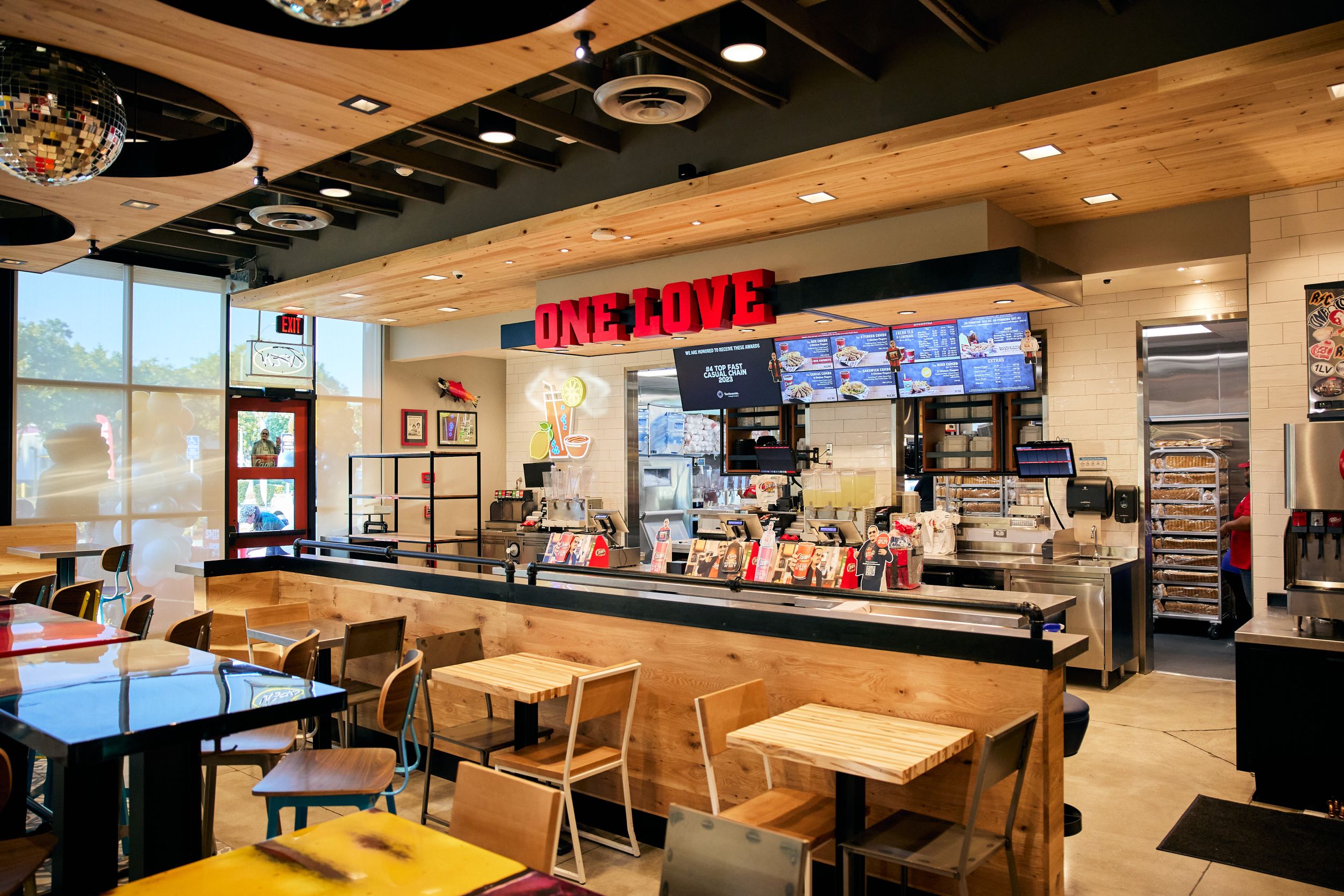 Interior view of a fast food restaurant showing a counter with digital menu boards, wooden tables and chairs in a modern setting.