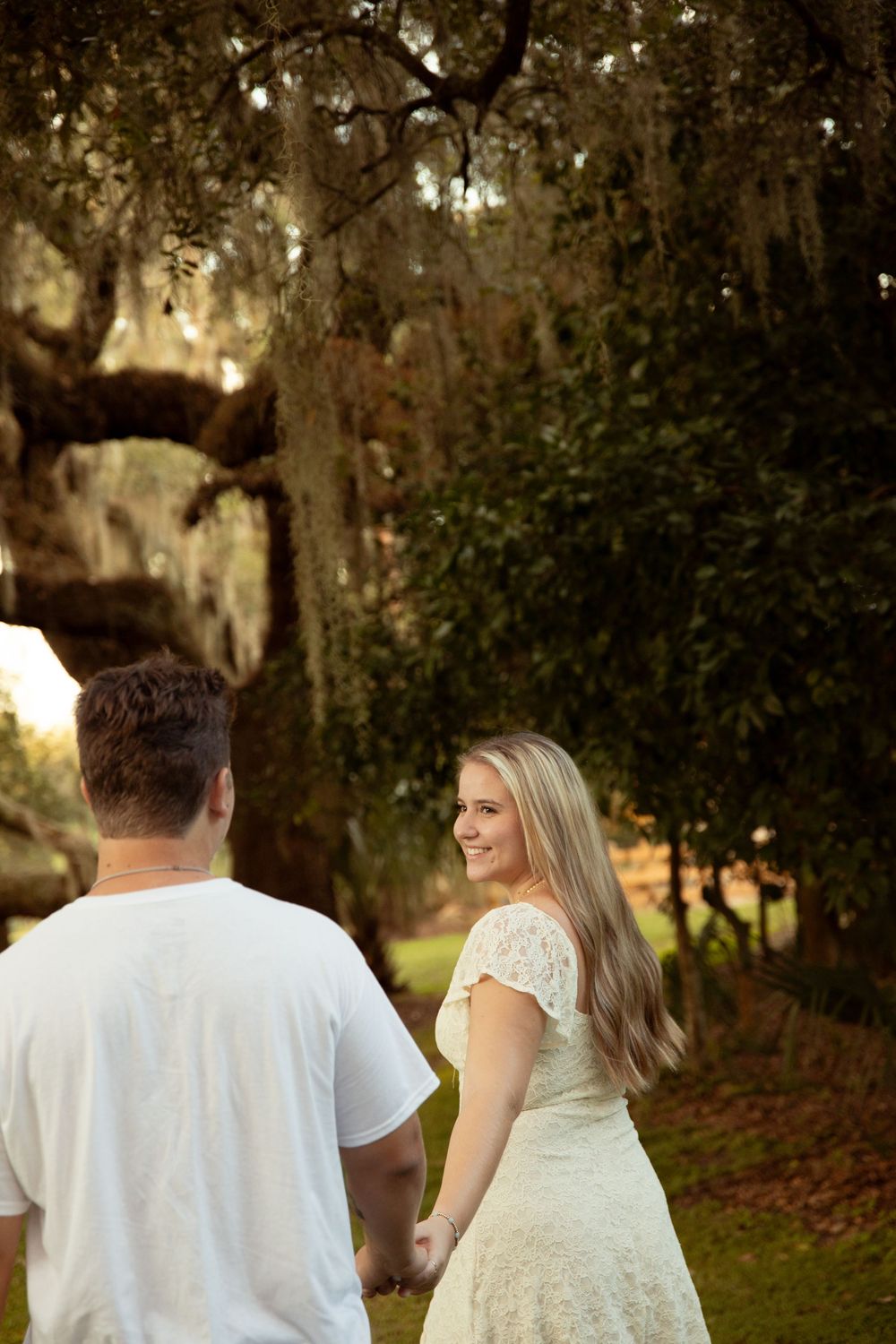 A couple in white attire walks together through a romantic garden setting at sunset.