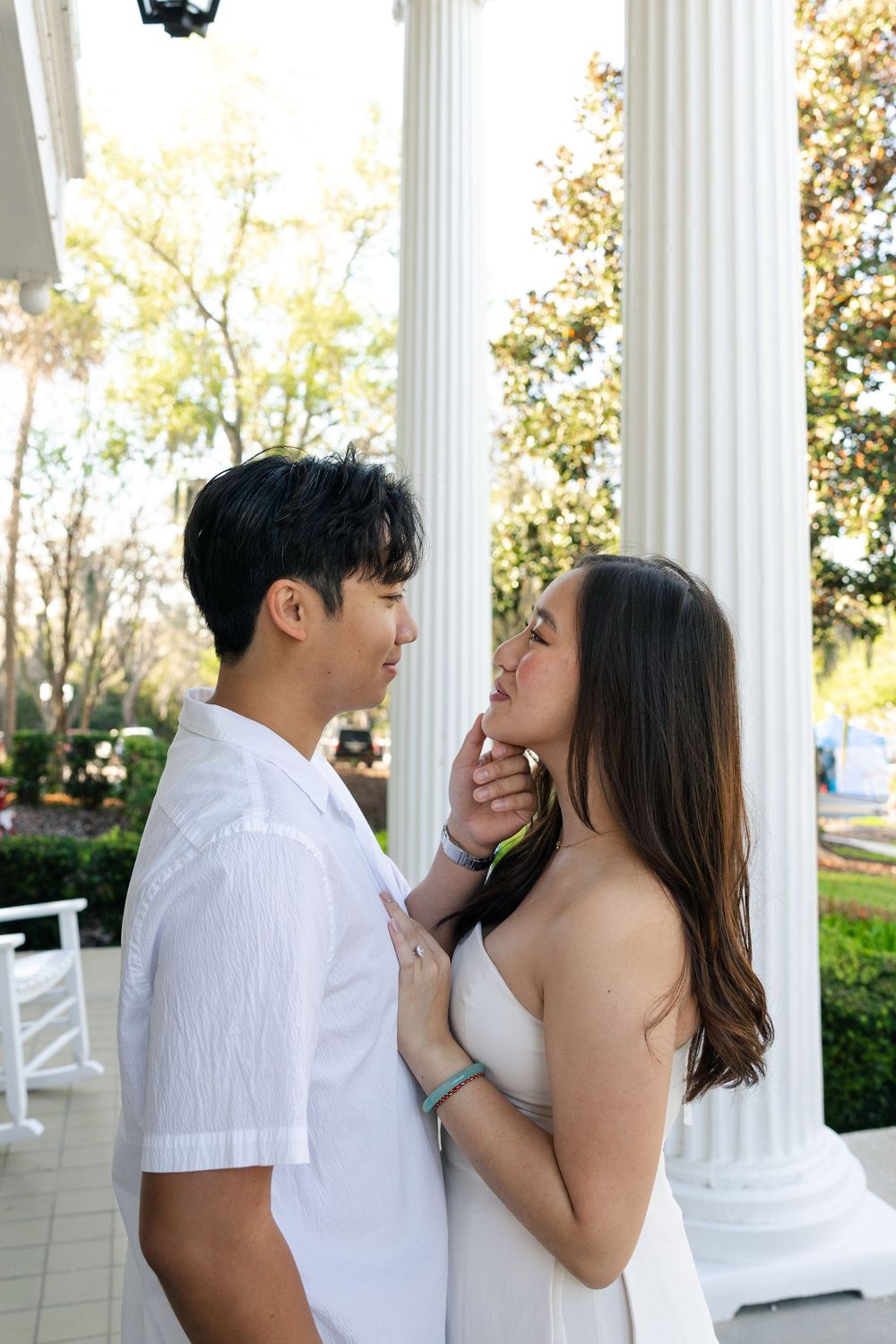 A romantic couple in white outfits share an intimate moment on a porch with white columns and greenery in the background.