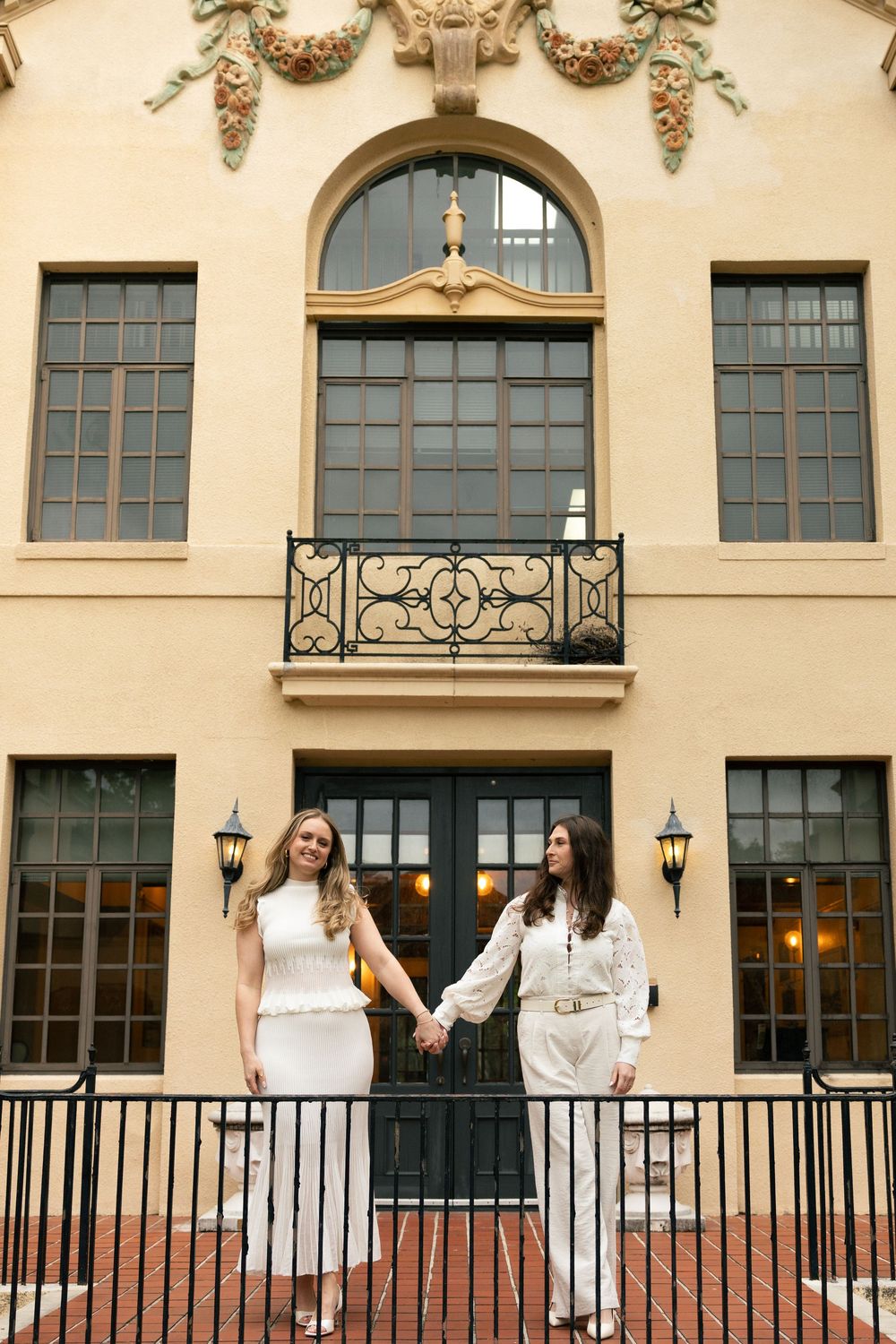 A group wearing white dresses stands together on a balcony of an elegant building with arched windows and ornate details.