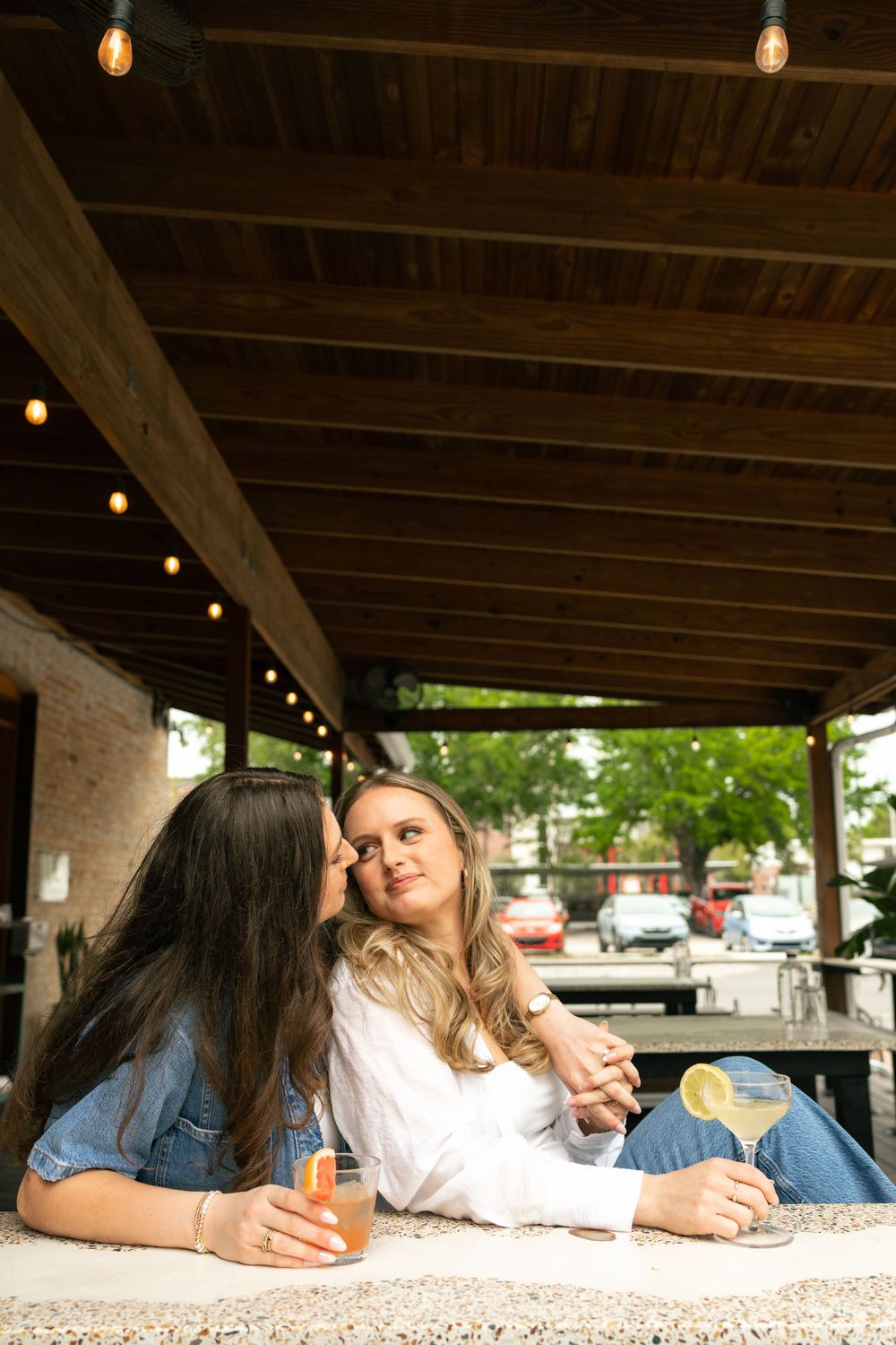 Two friends enjoy drinks together at an outdoor restaurant patio with wooden beams and hanging lights overhead.