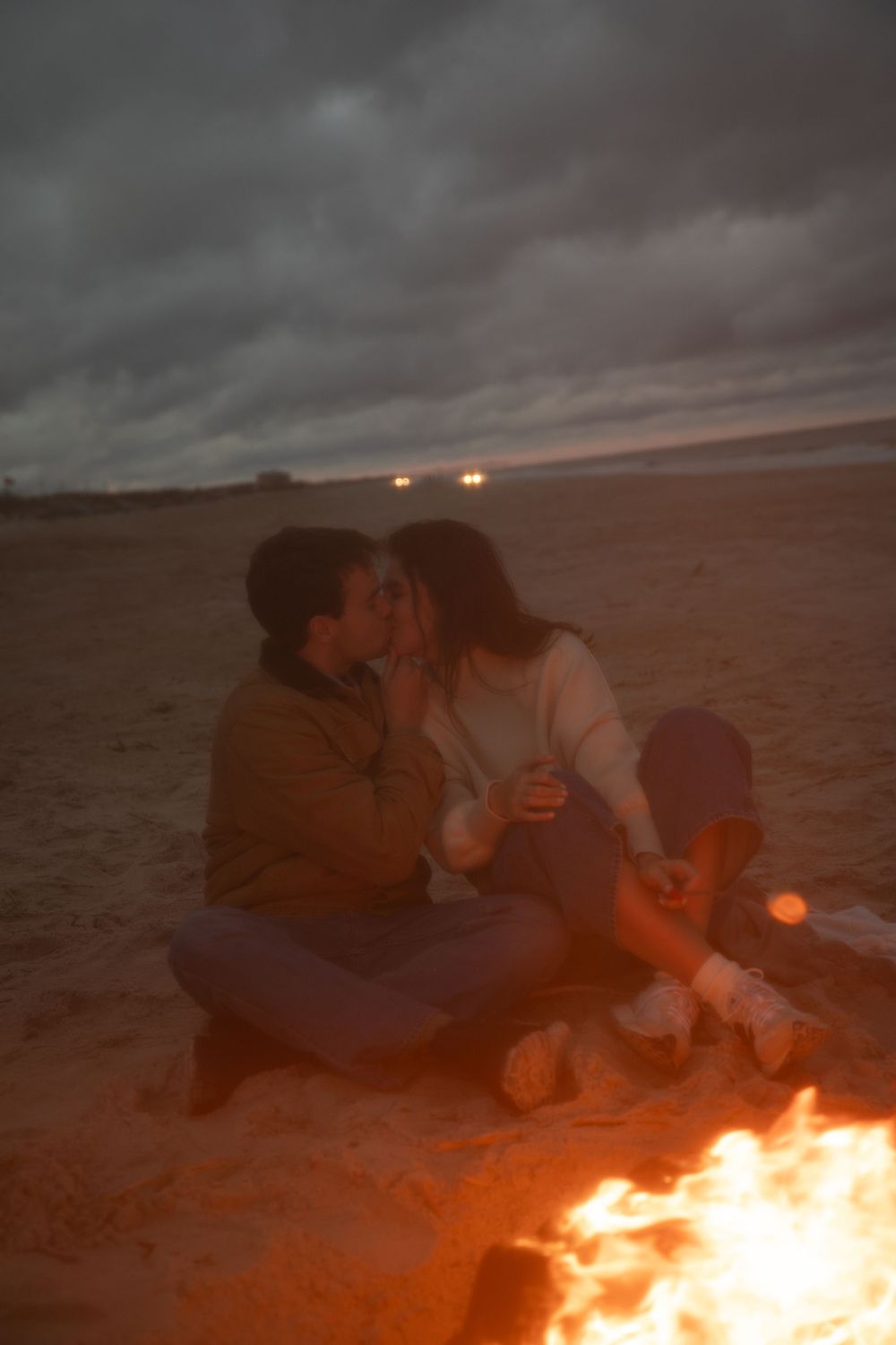 A couple gathered around a beach bonfire at night under stormy skies.