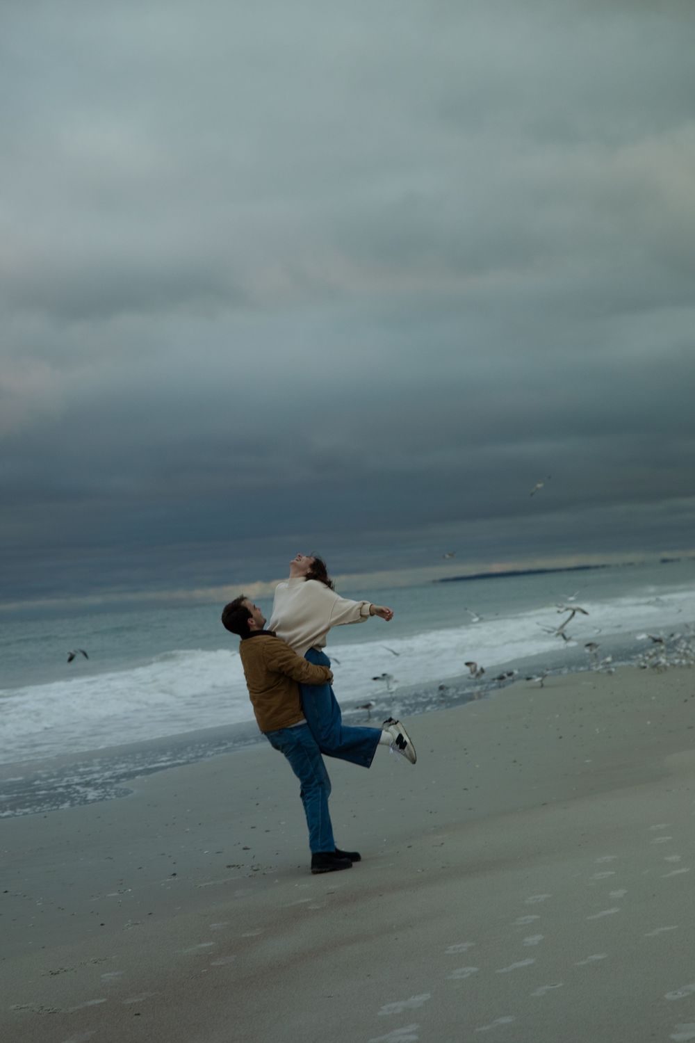 A couple in dramatic poses against moody beach landscape with stormy clouds.