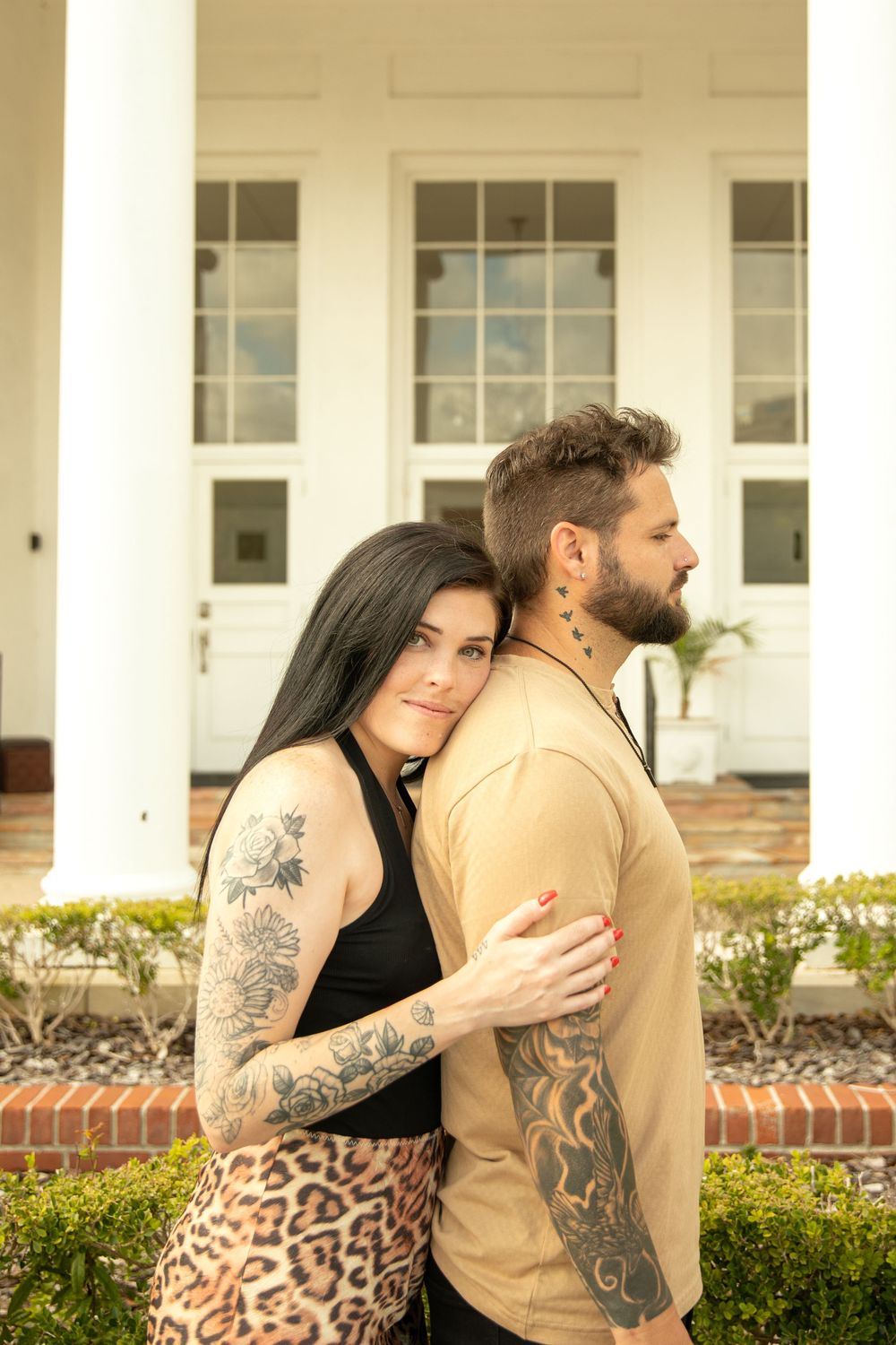 A tattooed couple poses intimately against white columns of a historic building.