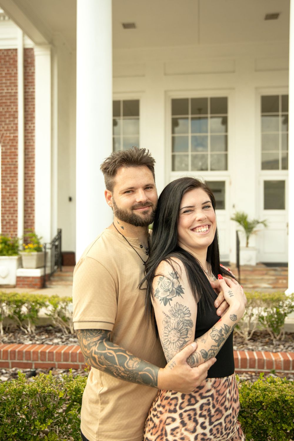 A happy couple with tattoos embraces while posing in front of a white columned building.