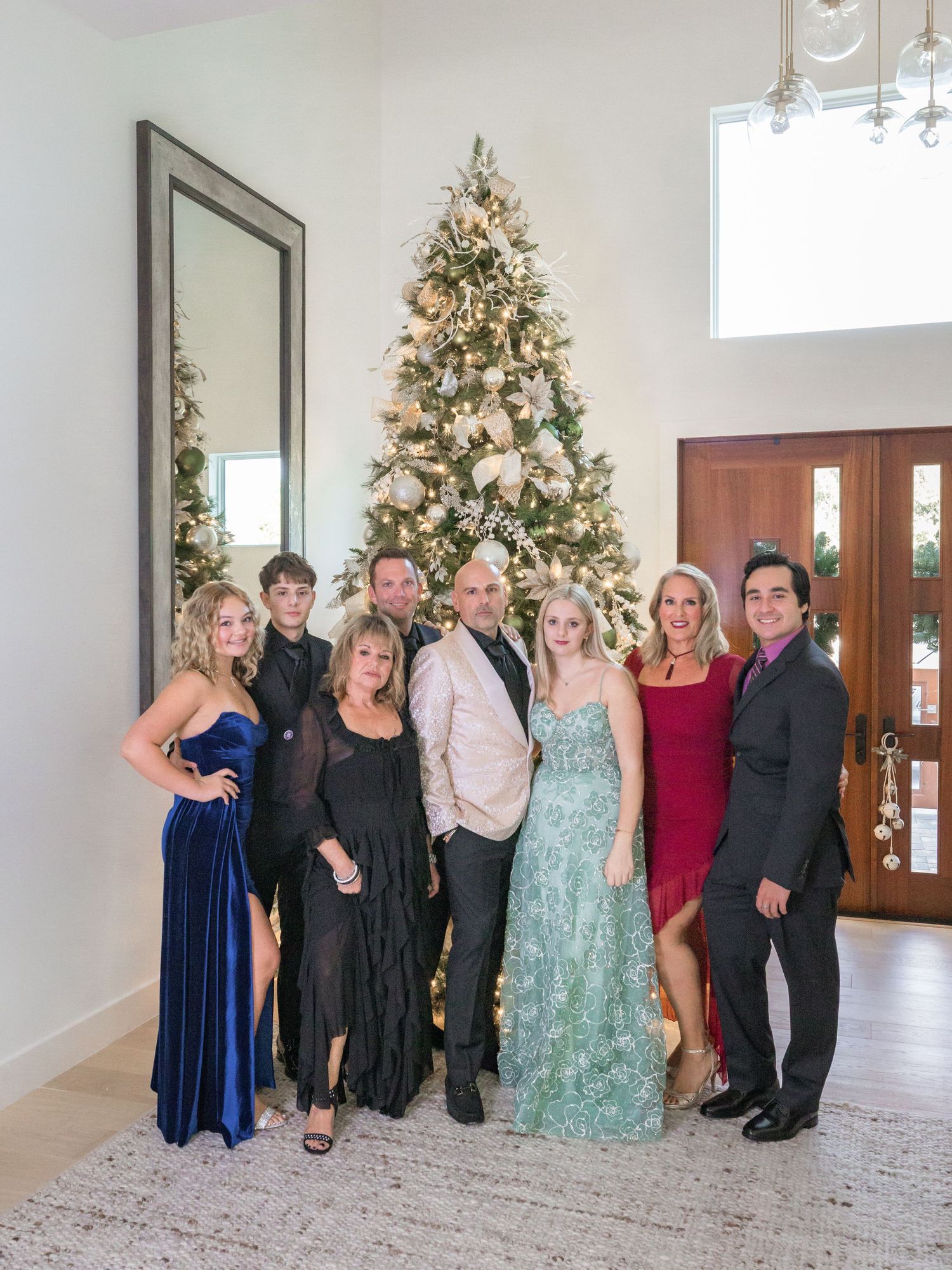 A group in formal attire poses together in front of decorated Christmas trees in an elegant white room.