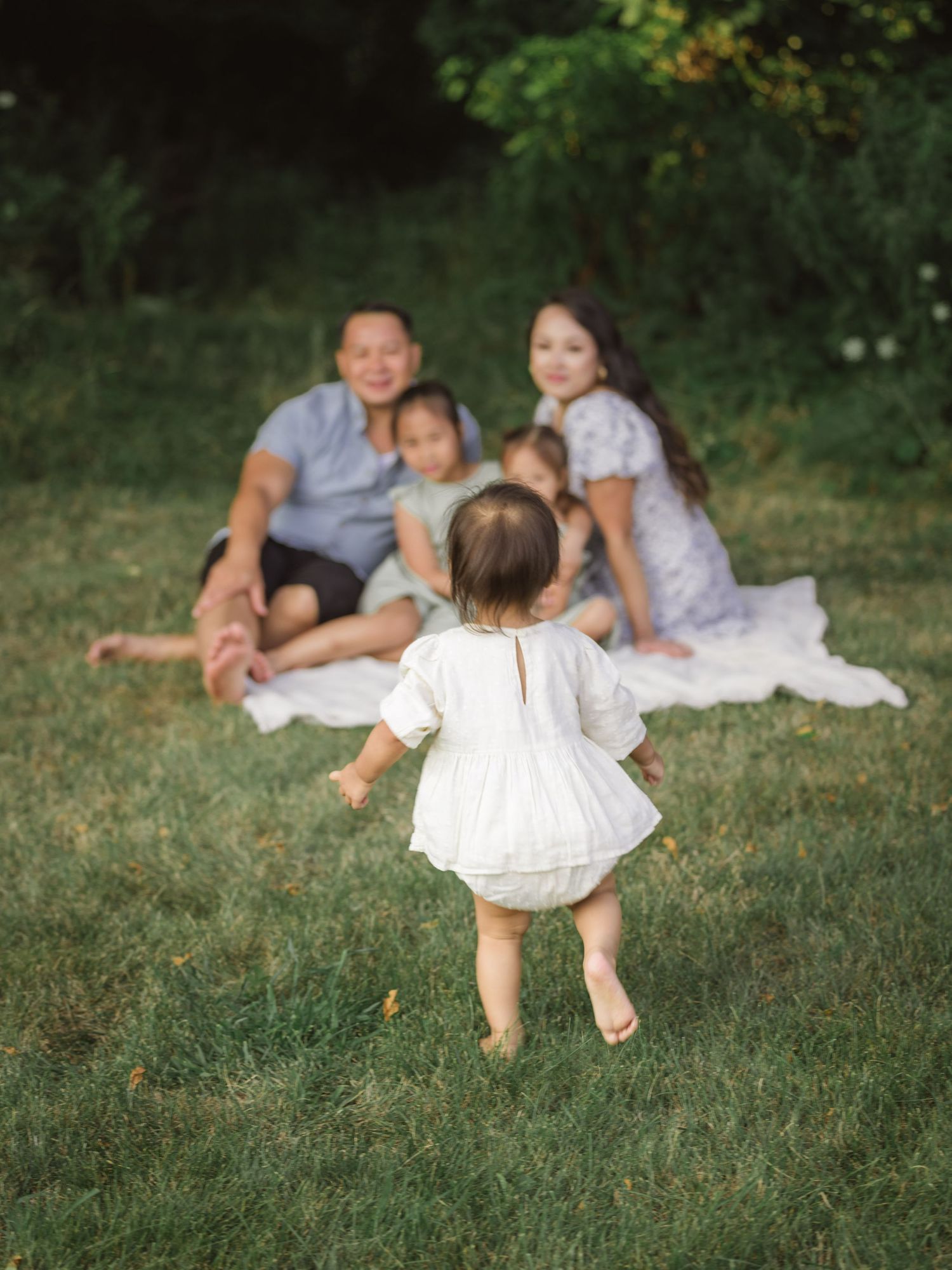 A toddler in a white dress runs across a grassy lawn toward a group sitting on a blanket outdoors.