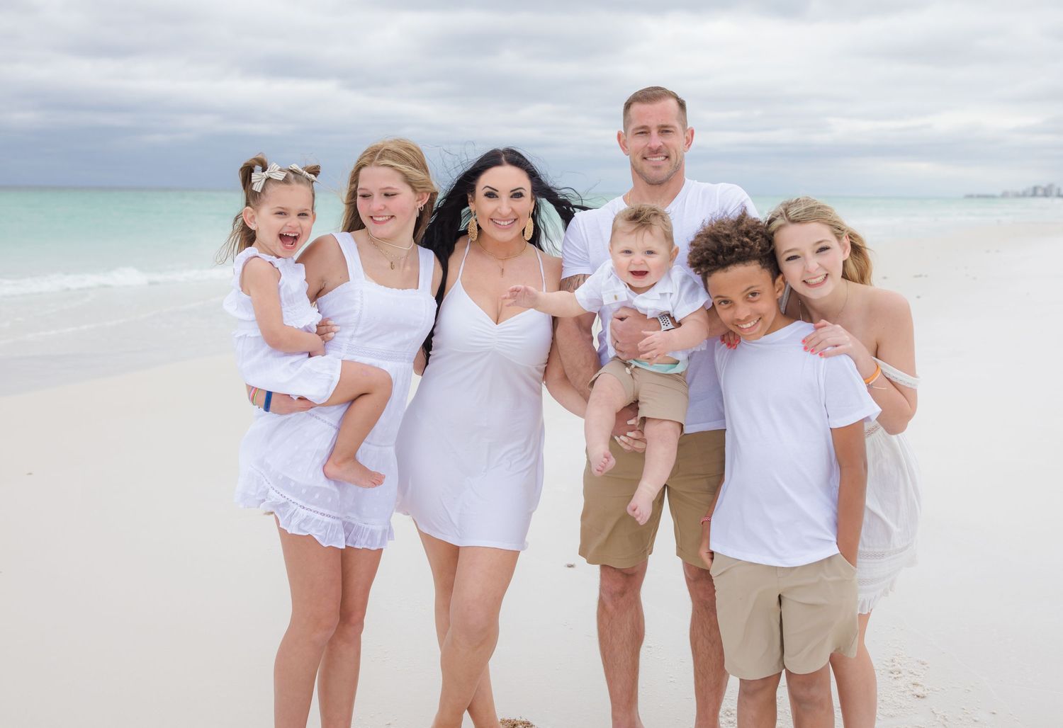 A family dressed in white and beige casual outfits poses together on a pristine beach with turquoise water and cloudy sky.