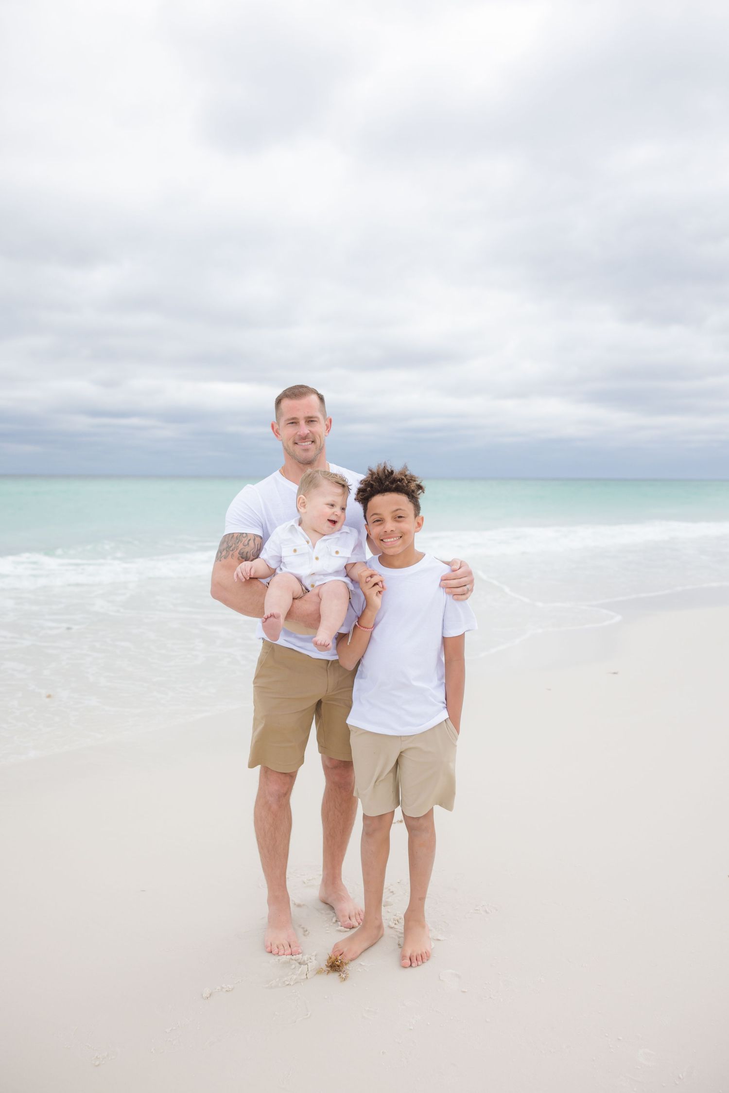 A joyful group stands together on a pristine white sandy beach with turquoise waters and cloudy skies in the background.