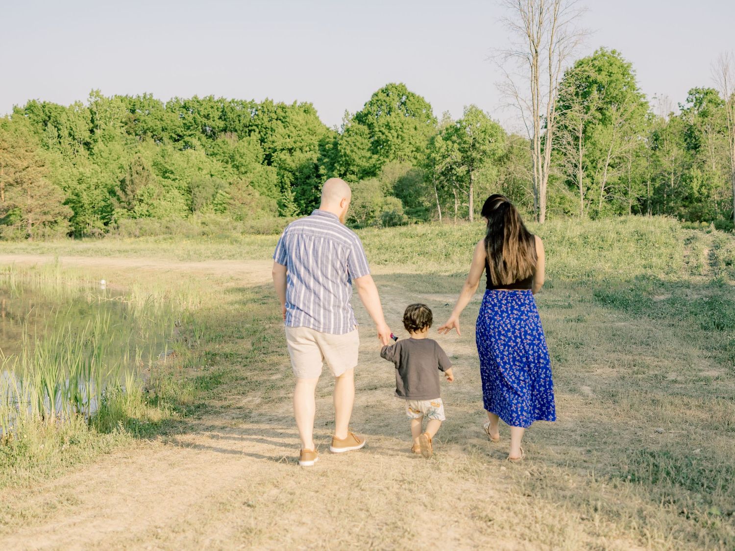 Three people walk hand in hand down a hillside path surrounded by lush summer greenery in a scenic outdoor setting.