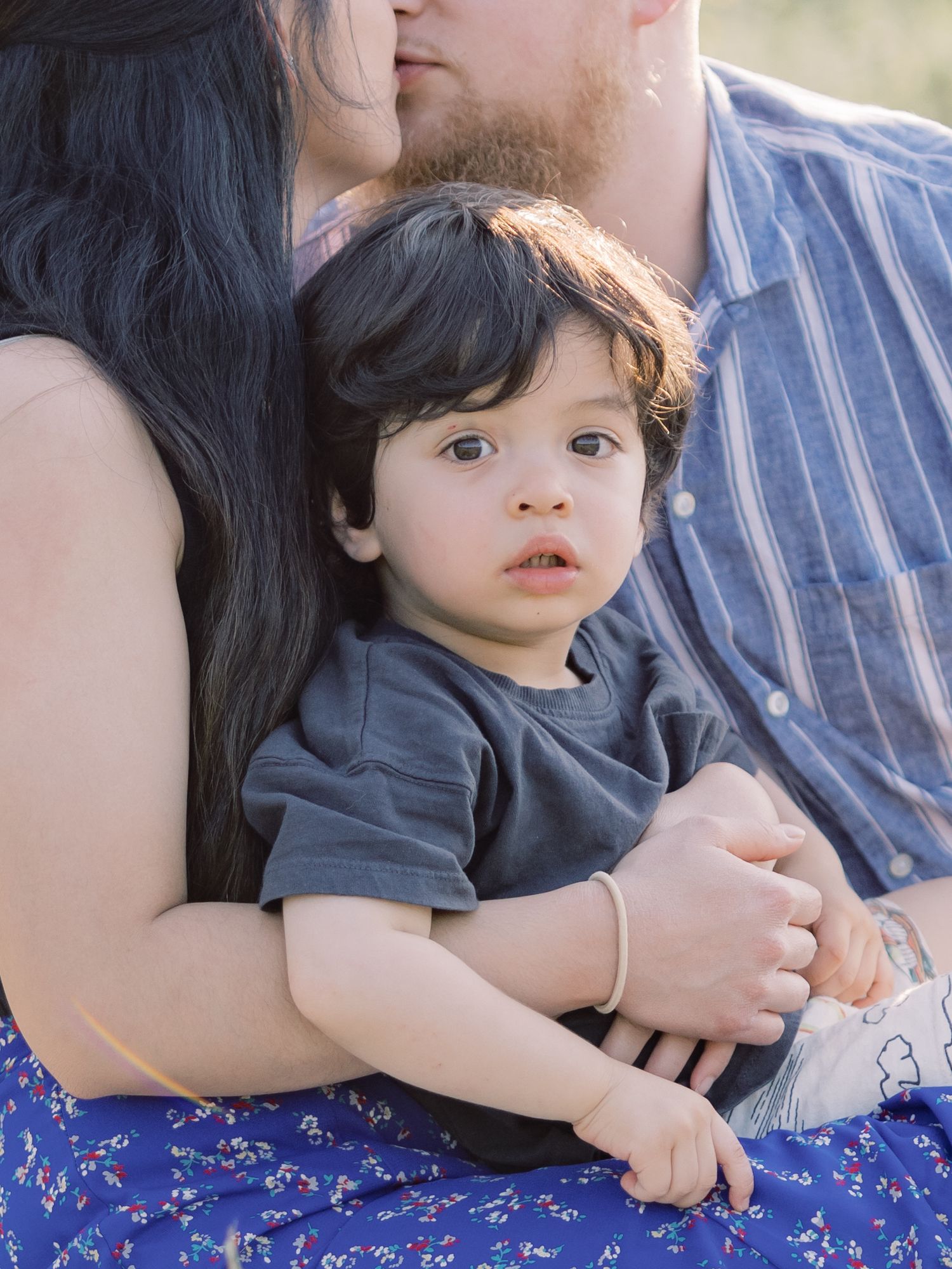 A toddler in a gray shirt sits between two adults on a blue blanket during an outdoor photo session.