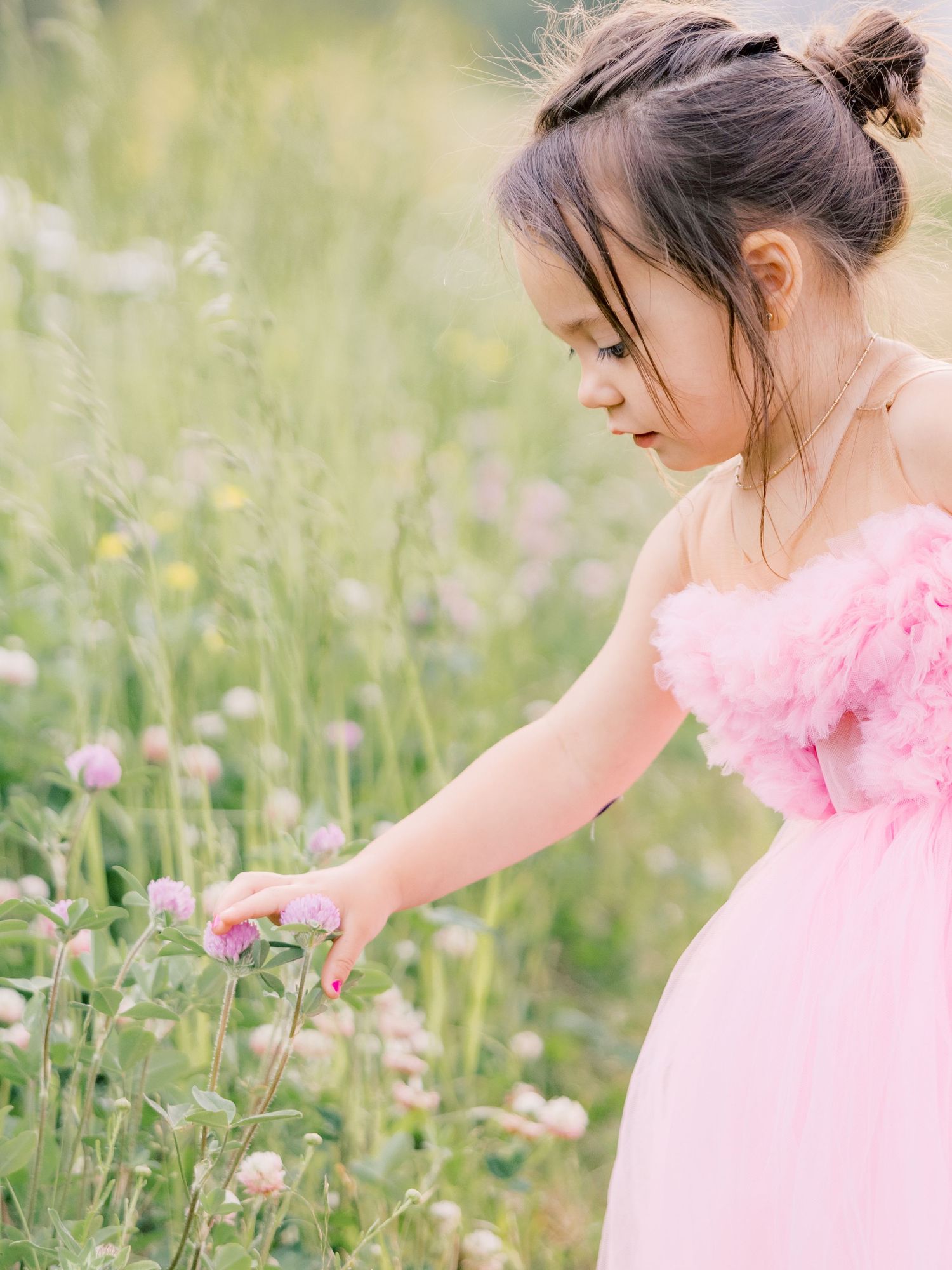 Young figure in a pink tulle dress gently reaches toward wildflowers in a dreamy summer meadow.