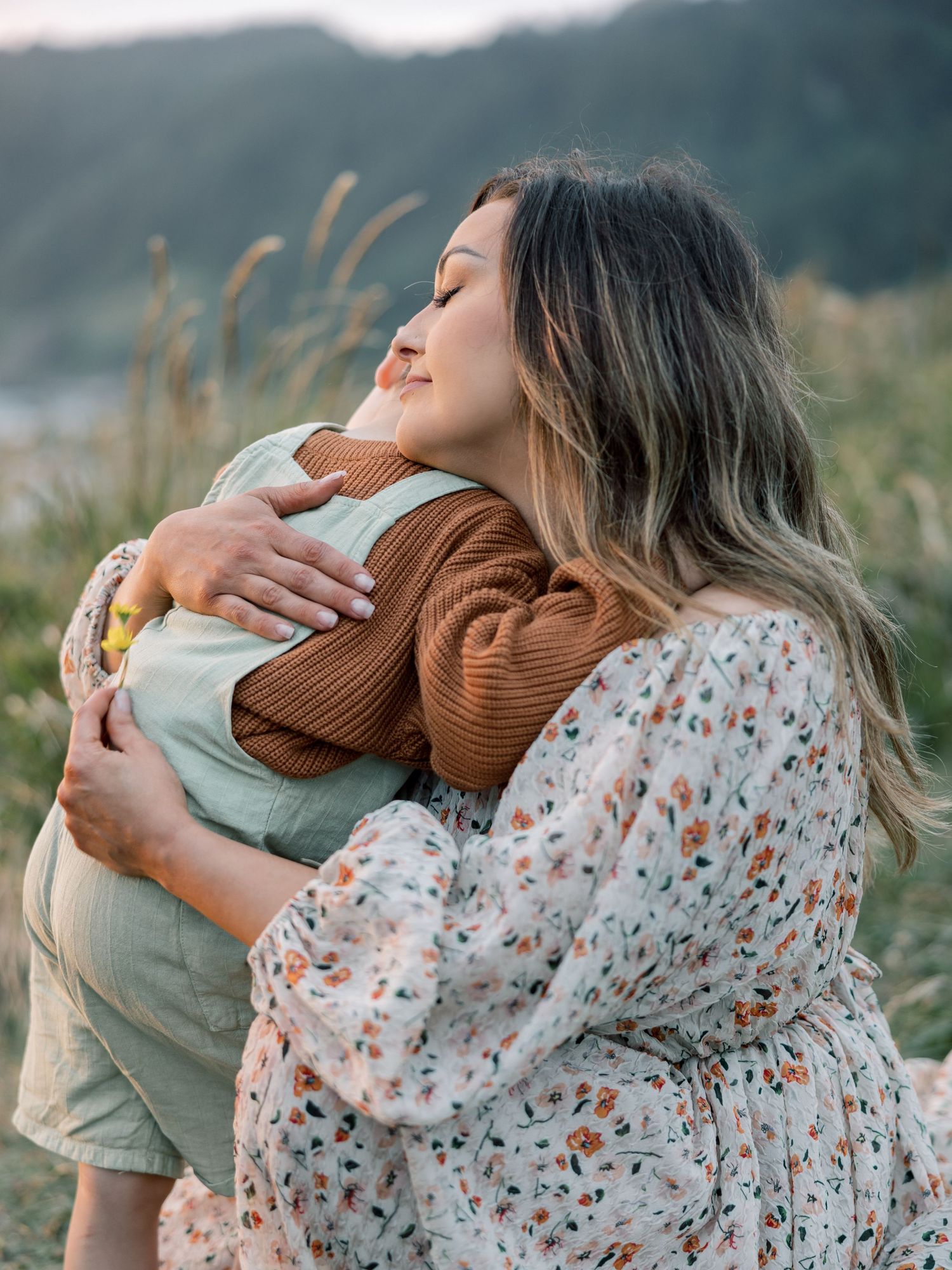 Tender embrace captured outdoors with warm brown sweater and floral dress against natural landscape background.