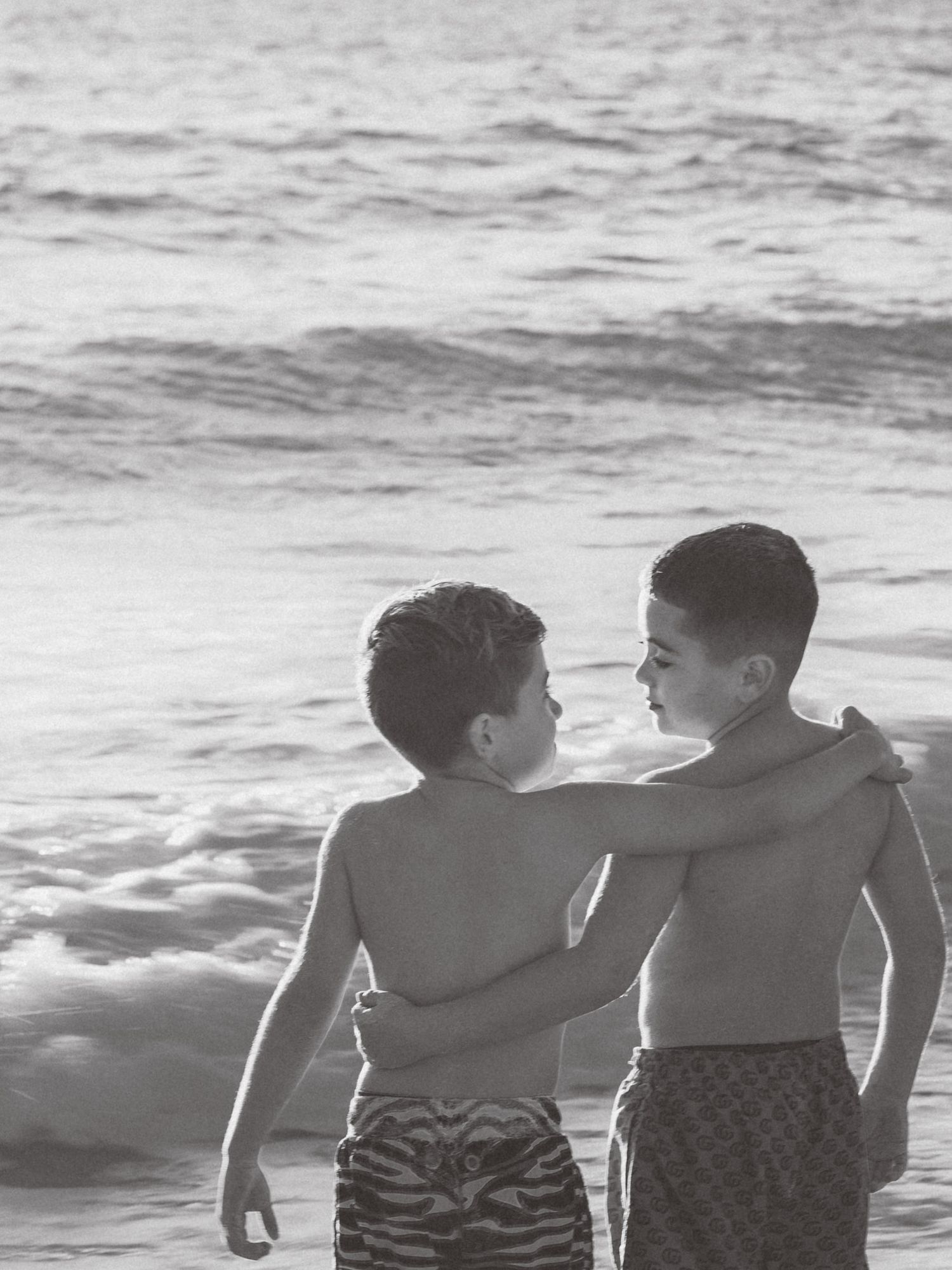 Two children stand close together at the beach in black and white, looking at each other with the ocean waves behind them.