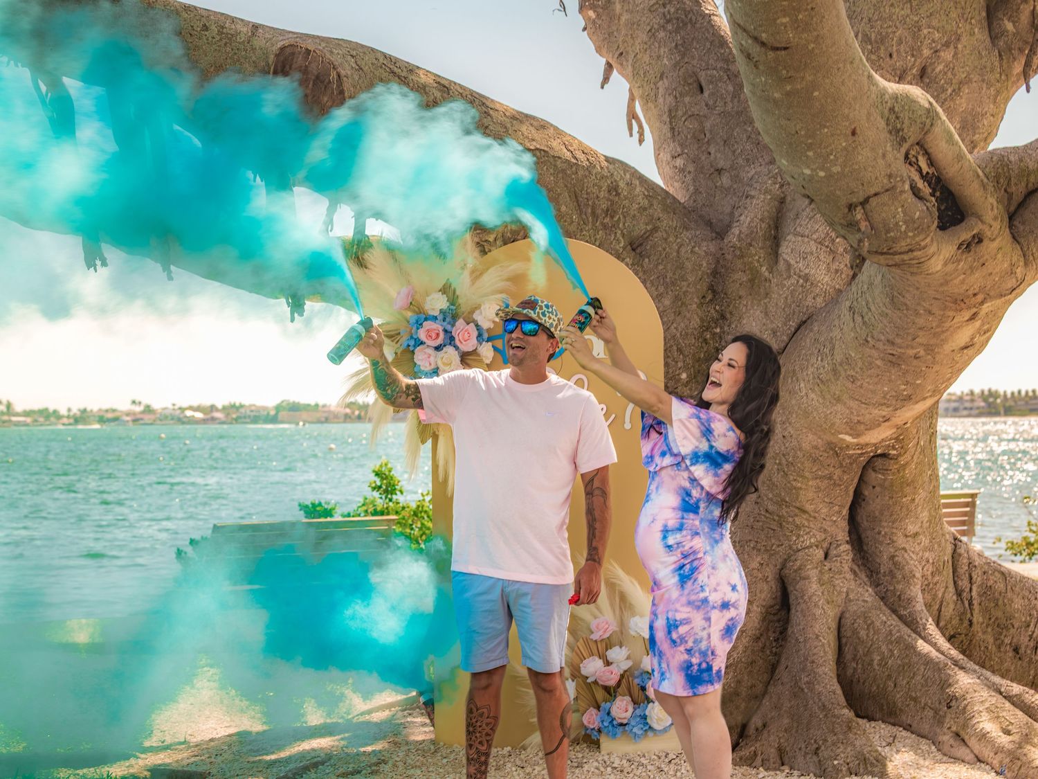 A family enjoys a scenic beach photoshoot under a large tree with turquoise ocean waters in the background.