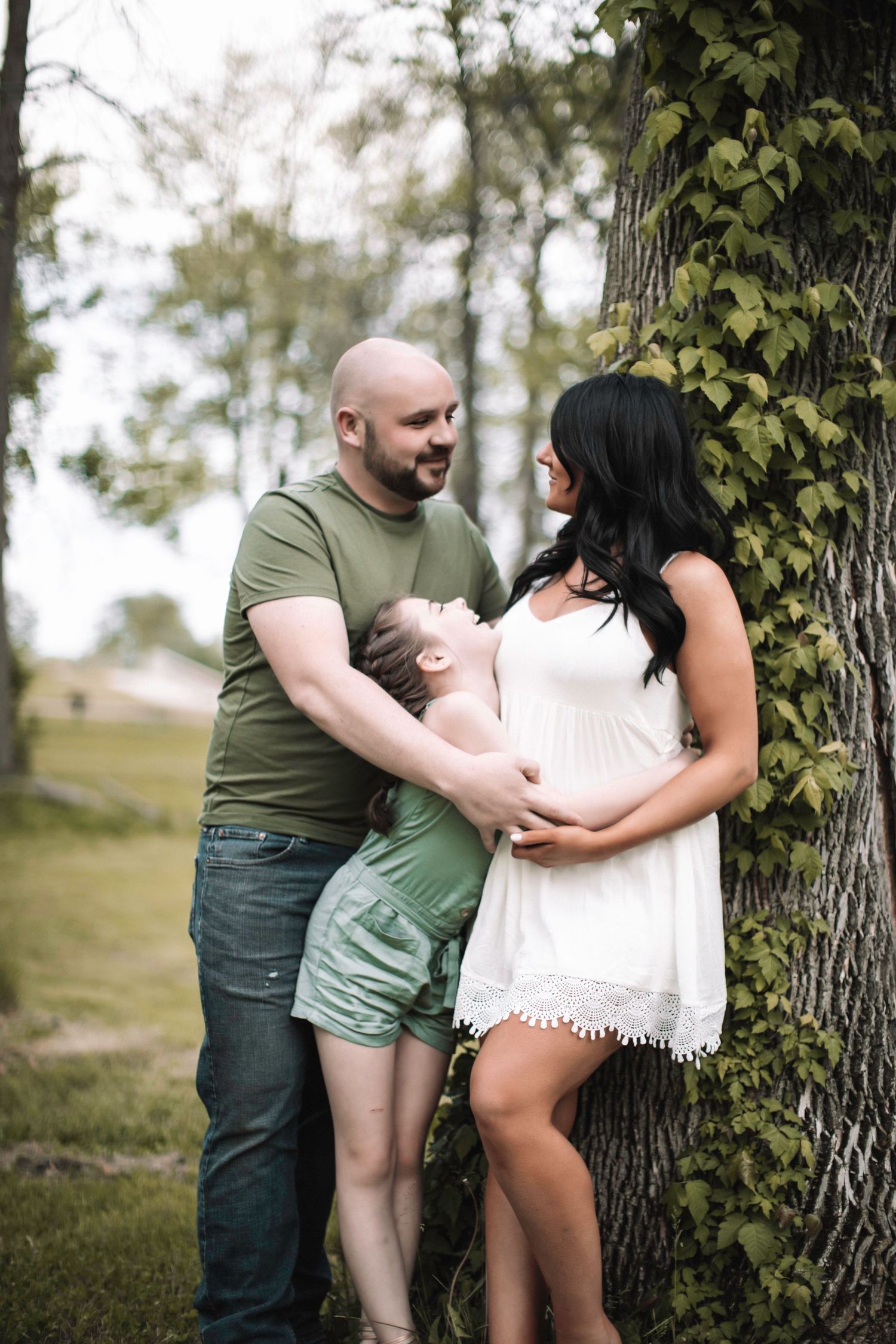 A couple and their child embrace while standing next to a tree in a park setting on a sunny day.