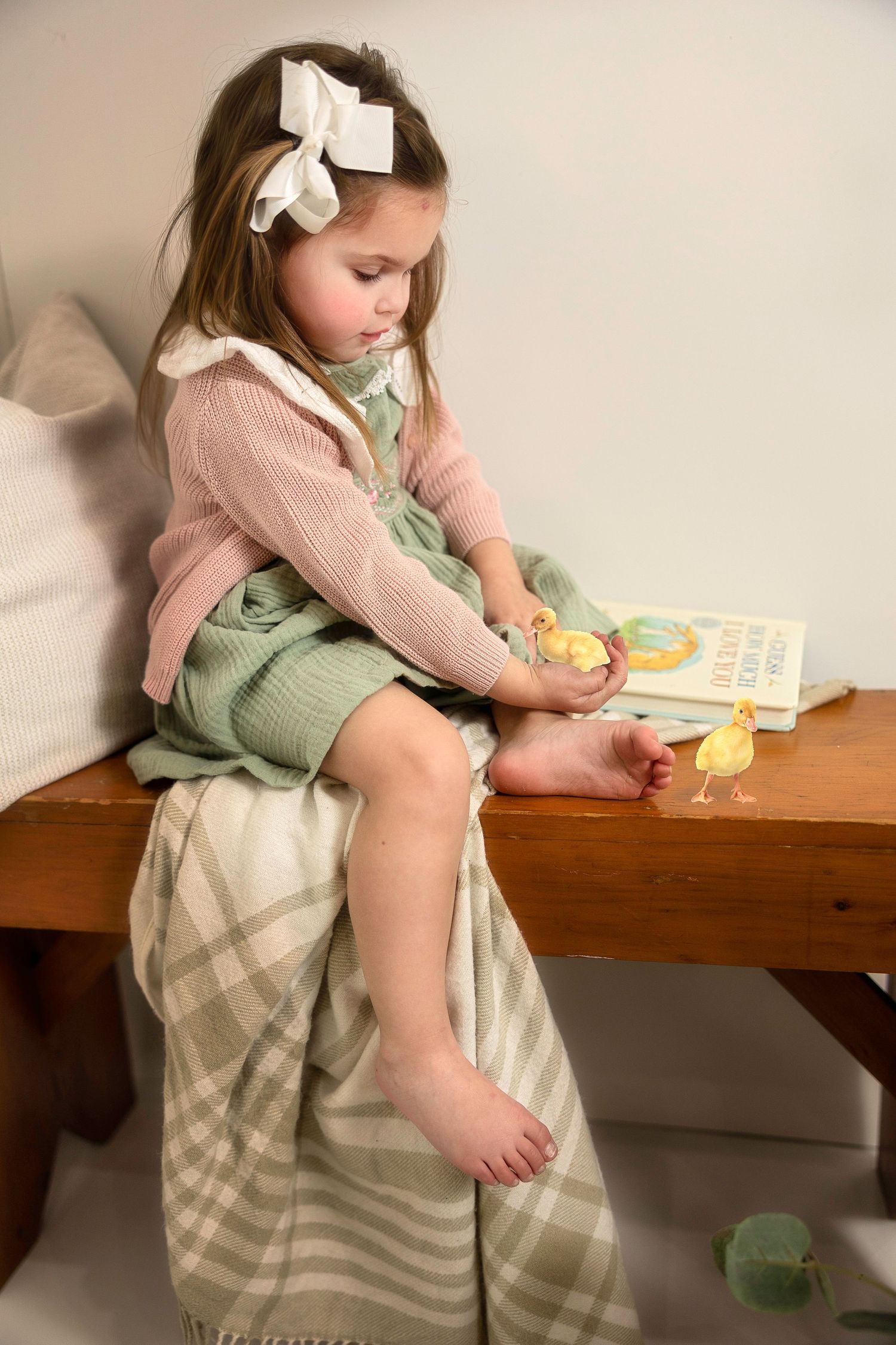 A child in a pink cardigan and sage green dress sits on a wooden surface while reading with a white bow in their hair.