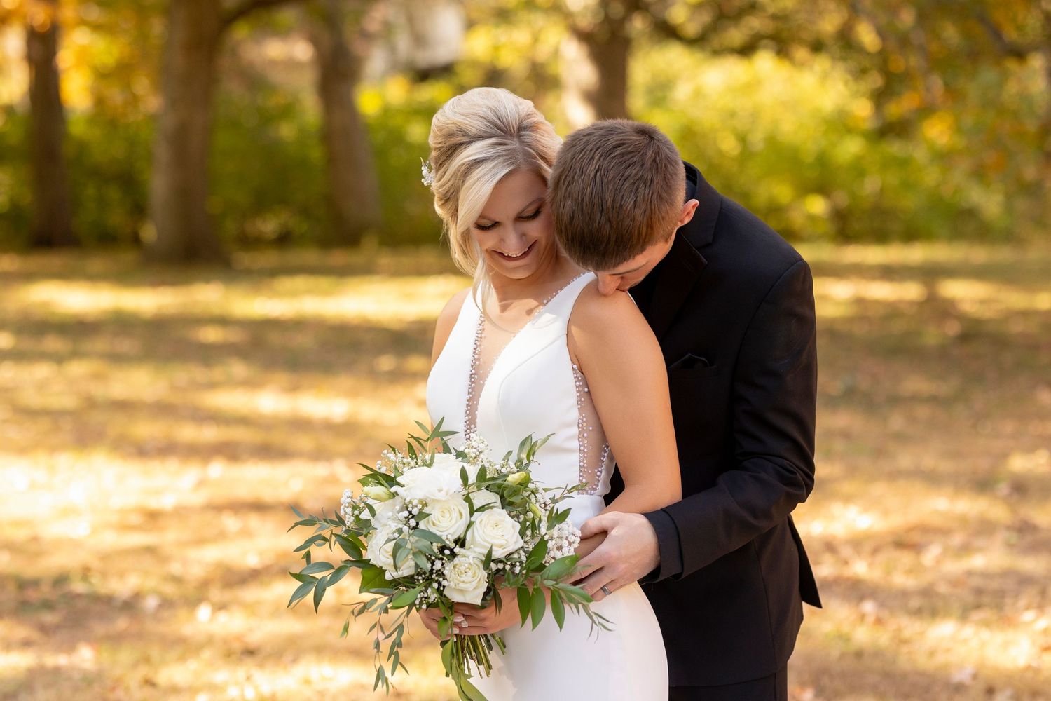 Romantic wedding couple embraces outdoors with white and green bouquet in autumn setting.