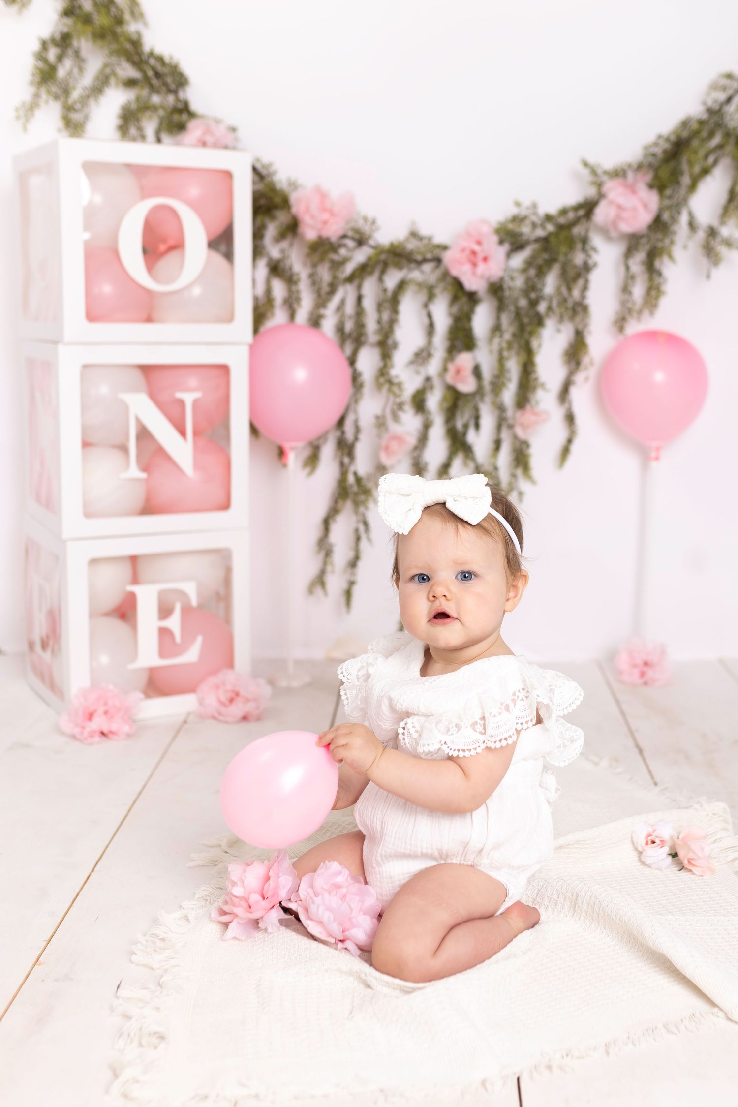 Pink and white first birthday photo setup features decorative ONE blocks, balloons, and evergreen garland accents.