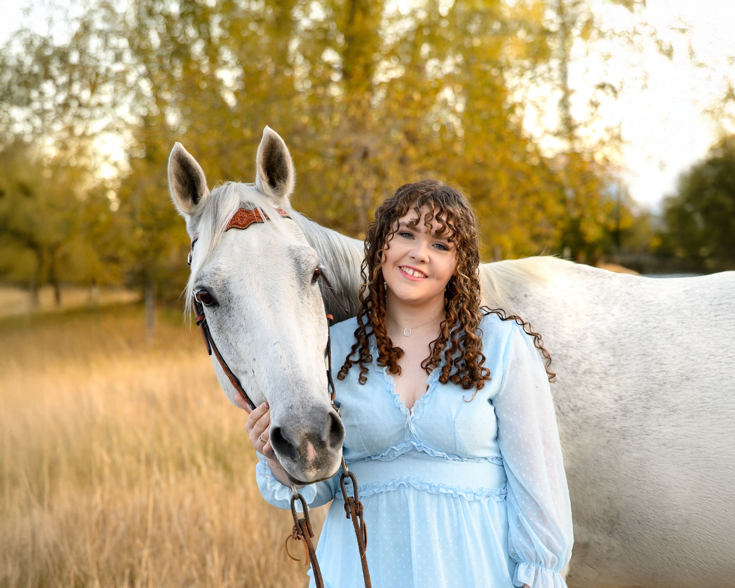 A gentle white horse and someone in a blue dress share a moment in an autumn field.