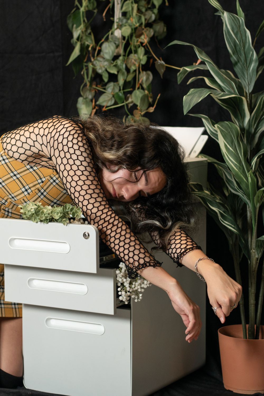 Person in fishnet top and plaid skirt reaches over white filing cabinets near potted plants against dark background.